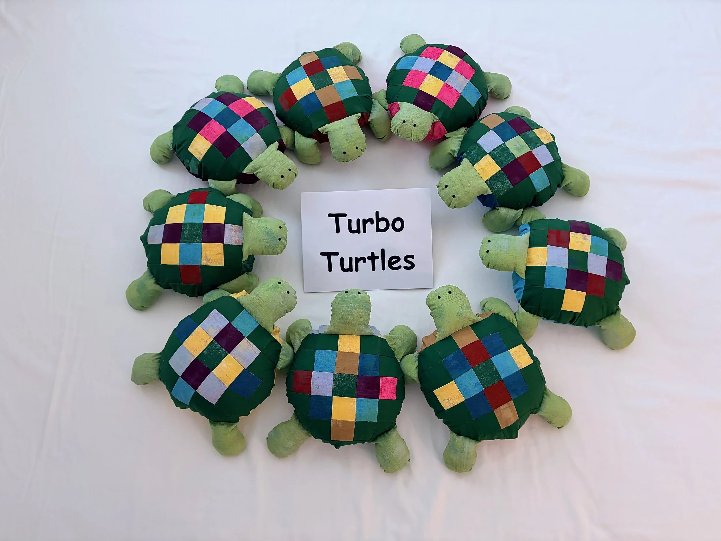 A group of colorful plush turtle toys with patchwork shells arranged on a white surface, with a sign reading "Turbo Turtles" in the center.