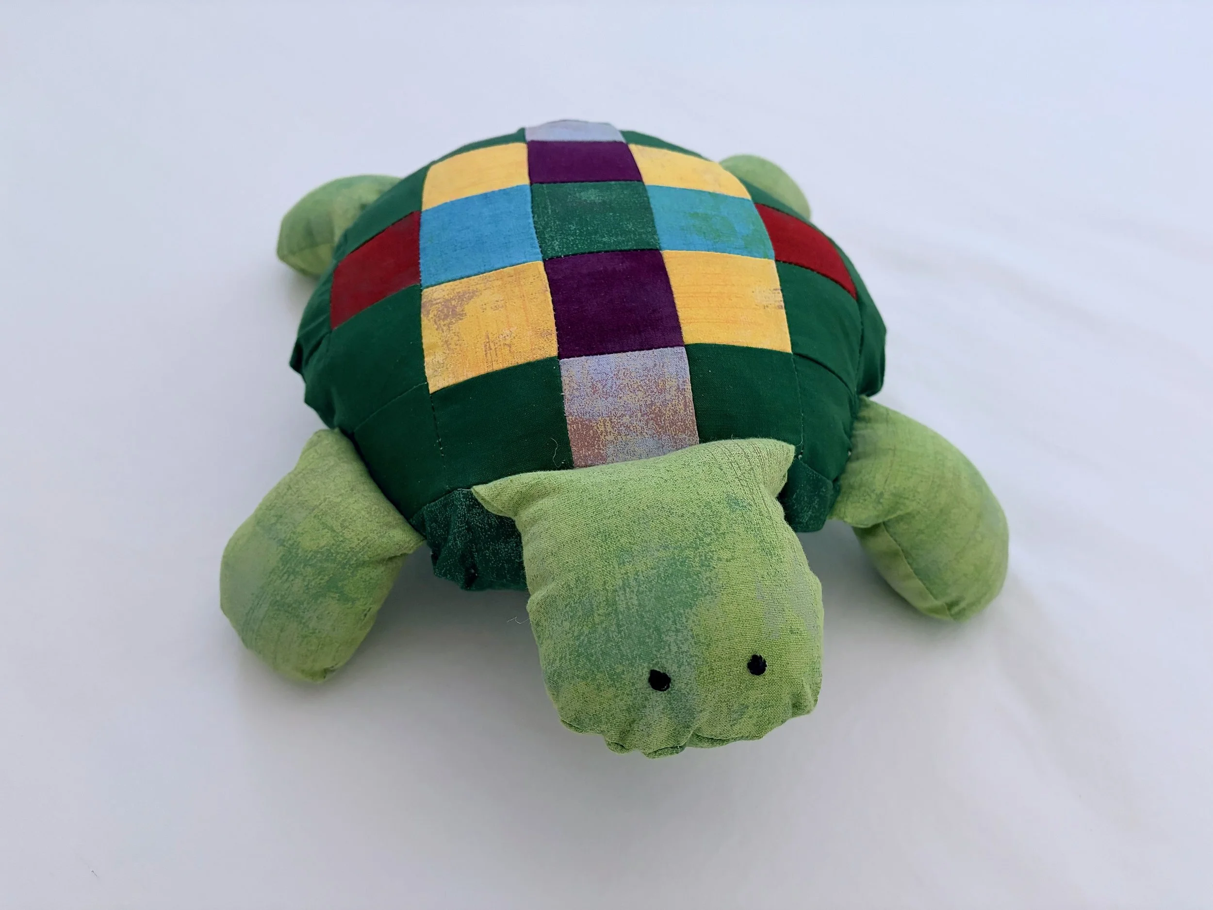 Colorful plush turtle toy with a patchwork shell featuring yellow, purple, blue, and red squares, green head and limbs, and small black eyes, set against a plain white background.