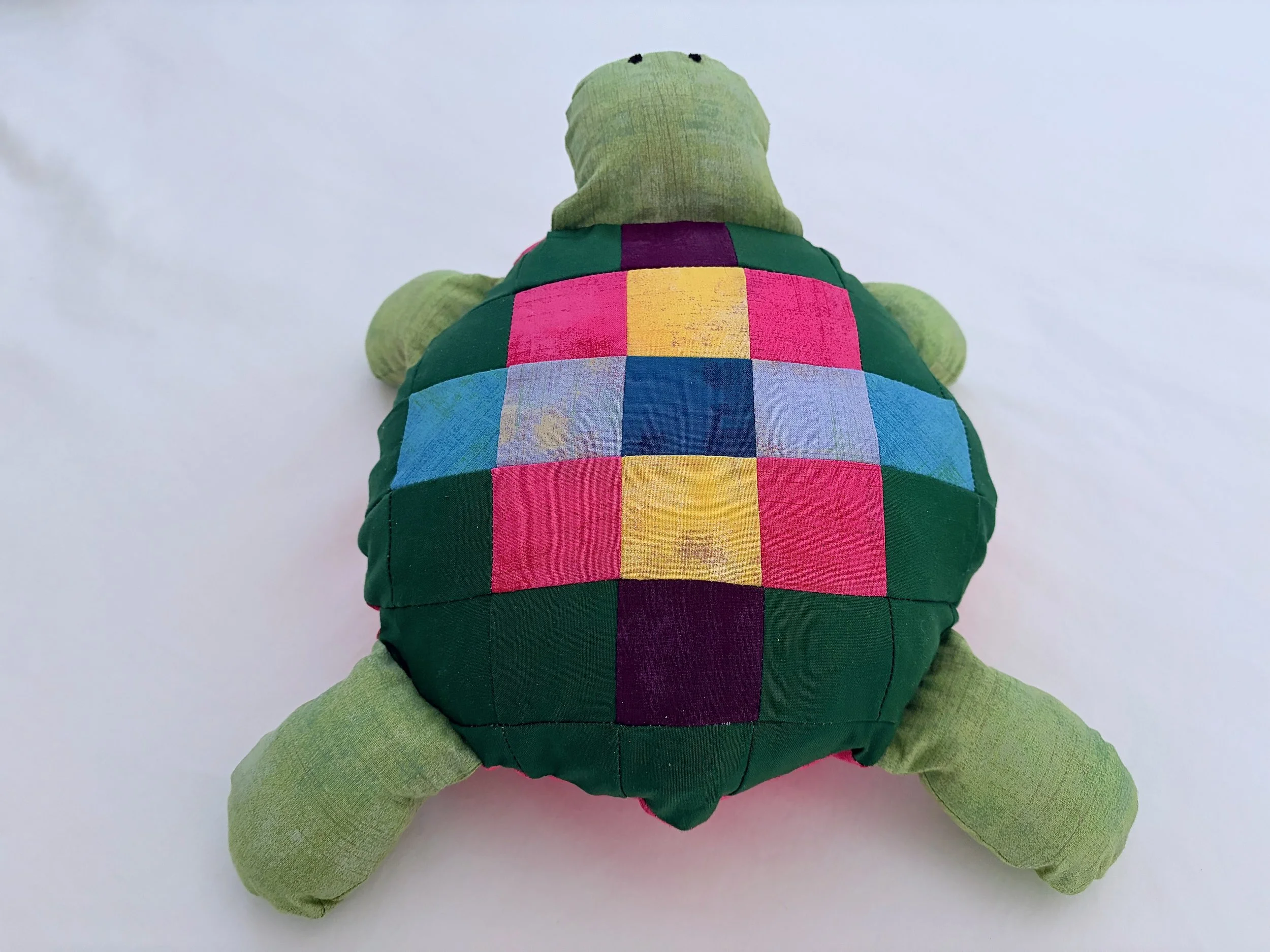 A green plush turtle toy with a multicolored patchwork shell, viewed from the back against a light background.
