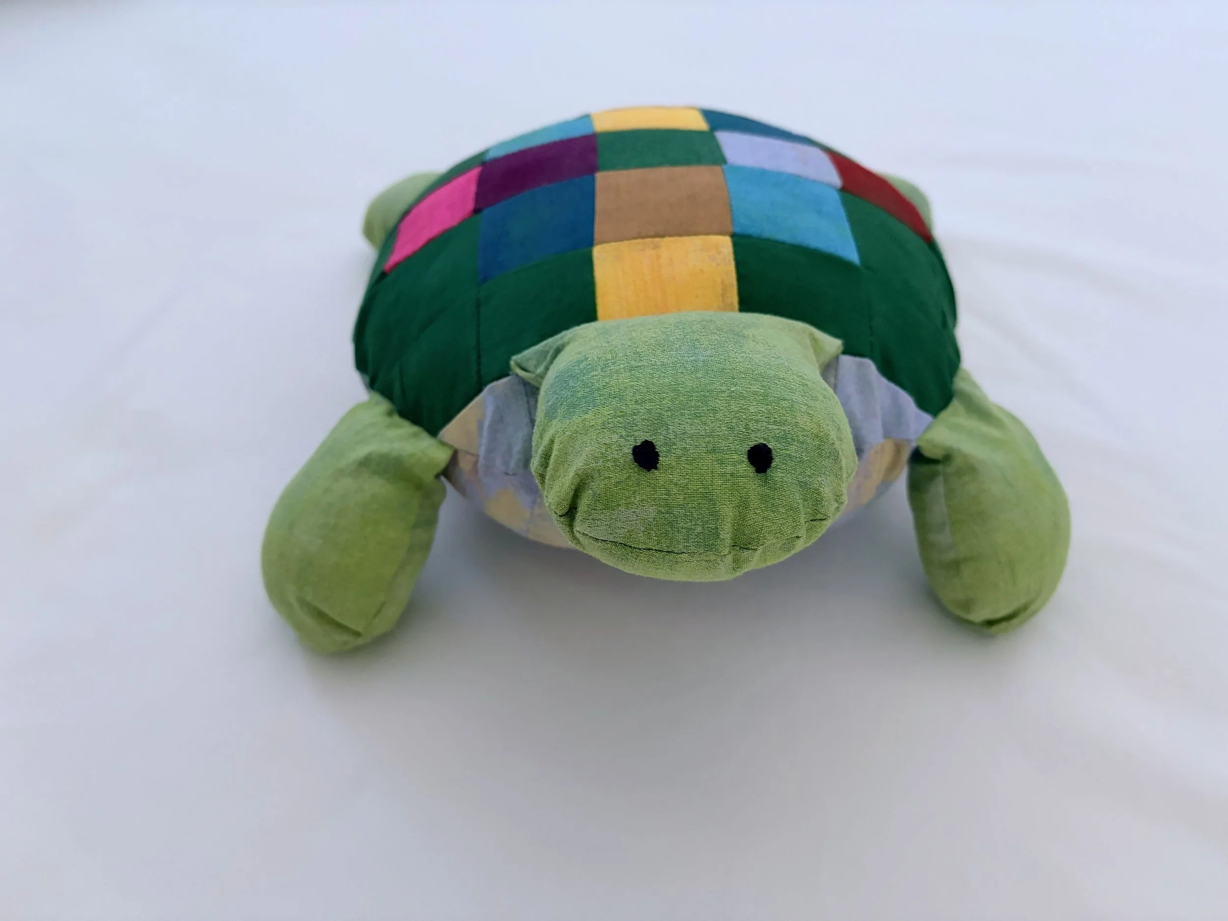 A stuffed turtle toy with a green body and a colorful patchwork shell, placed on a white surface.