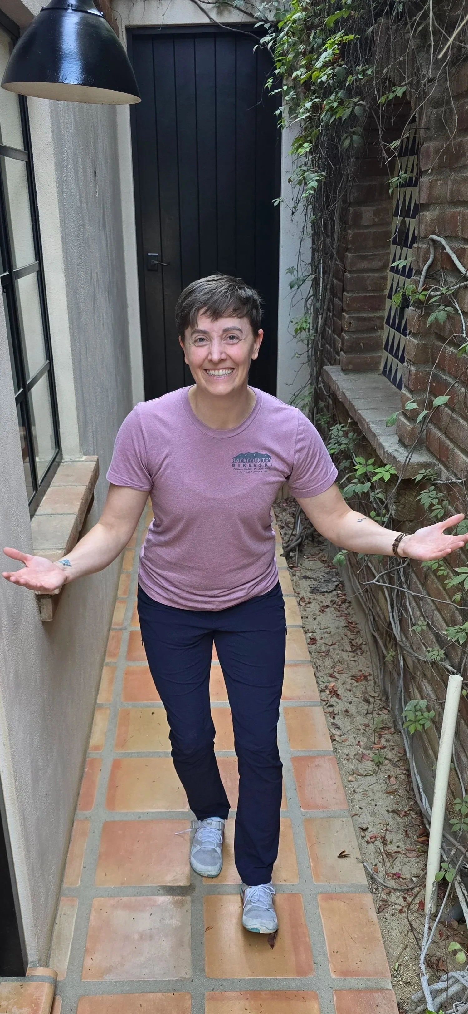 A smiling woman with short brown hair, wearing a pink T-shirt and navy pants, walking in a narrow outdoor pathway with terracotta tiles, surrounded by brick and stucco walls with some green vines.
