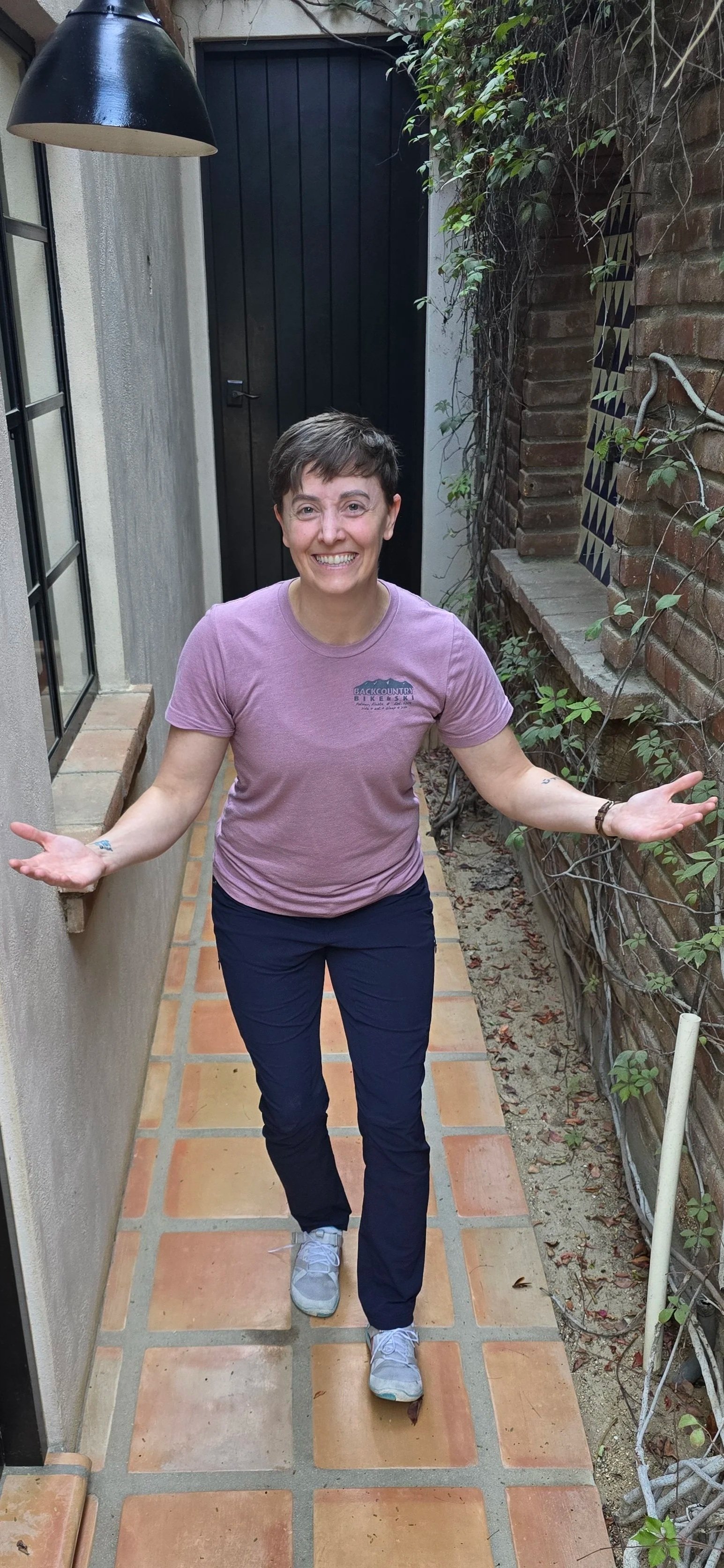 A woman with short brown hair wearing a pink T-shirt, black pants, and gray sneakers standing on a tiled walkway in a narrow outdoor space. She has a cheerful expression with her arms open widely. The background features a black wooden door, a beige 