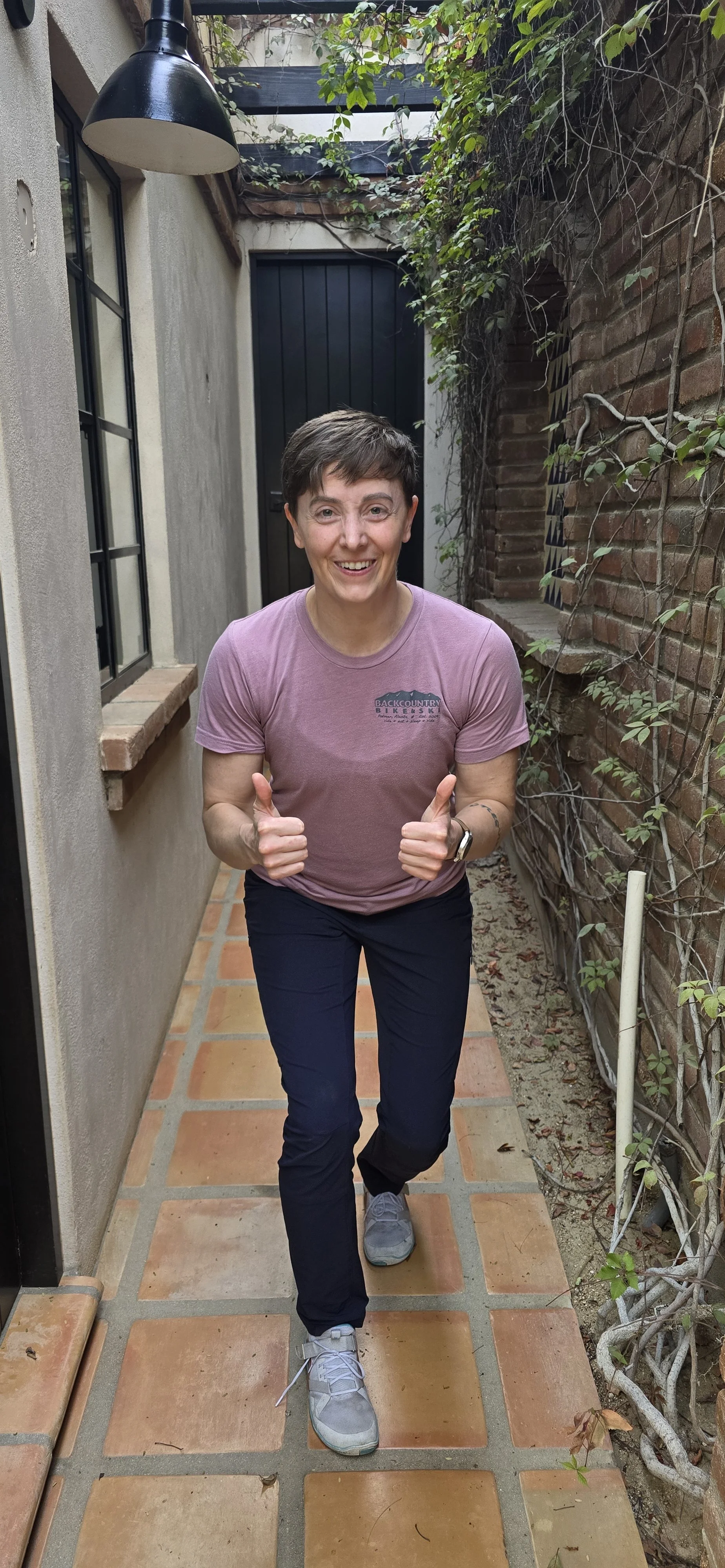A person with short brown hair smiling and giving two thumbs up, walking on a narrow brick patio between a beige stucco wall and a brick wall with climbing plants, wearing a purple t-shirt, black pants, gray running shoes, and a smartwatch.