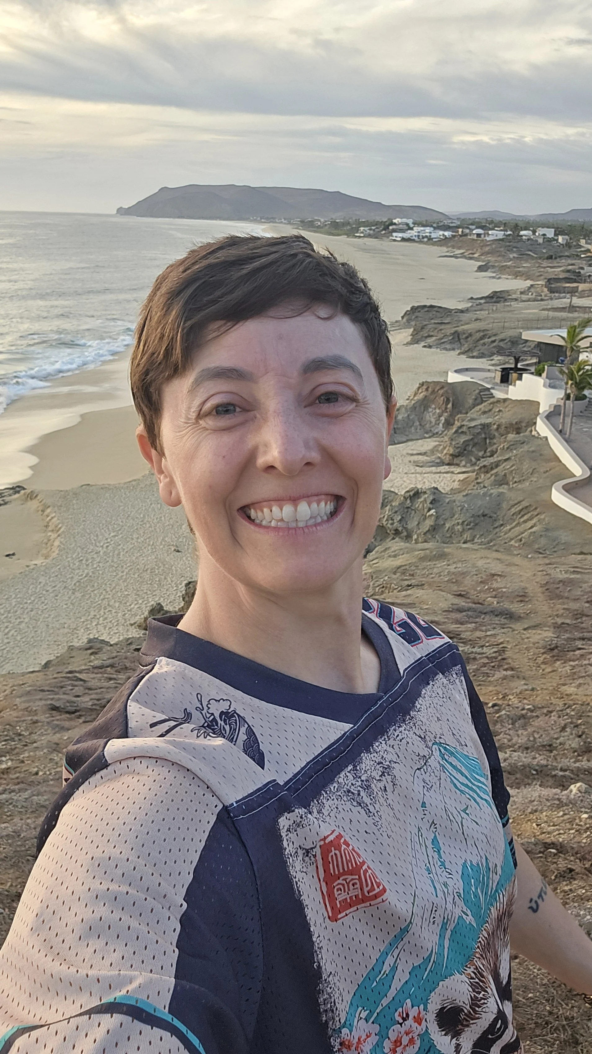 A person smiling on a rocky coastal area with sandy beach, ocean waves, distant mountains, and residential buildings in the background during sunset.