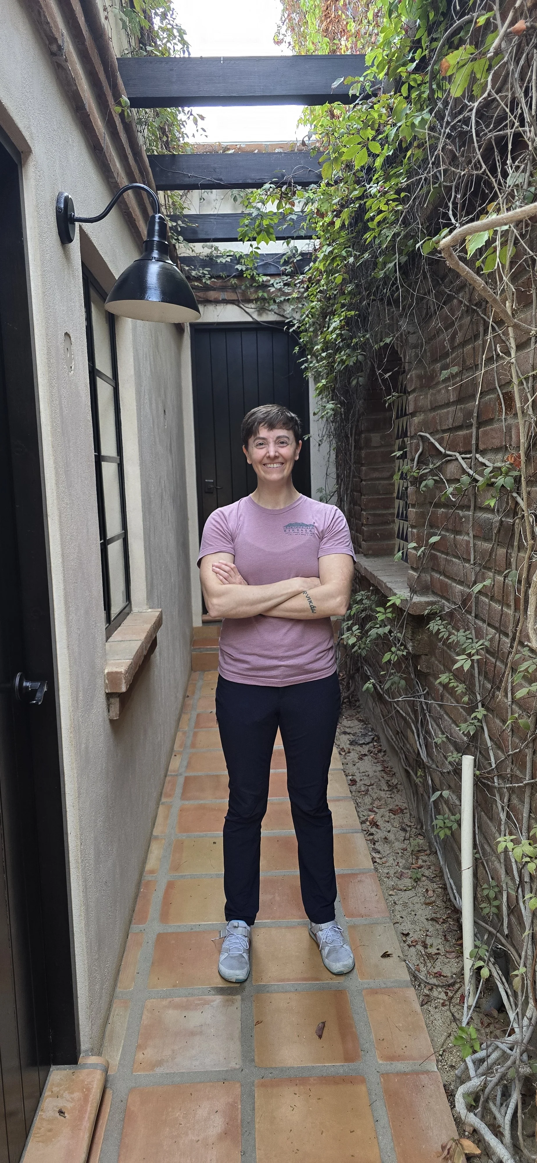 A woman with short dark hair, wearing a pink T-shirt and black pants, standing with arms crossed in a narrow outdoor corridor with brick and stucco walls, green vines, and a black door.