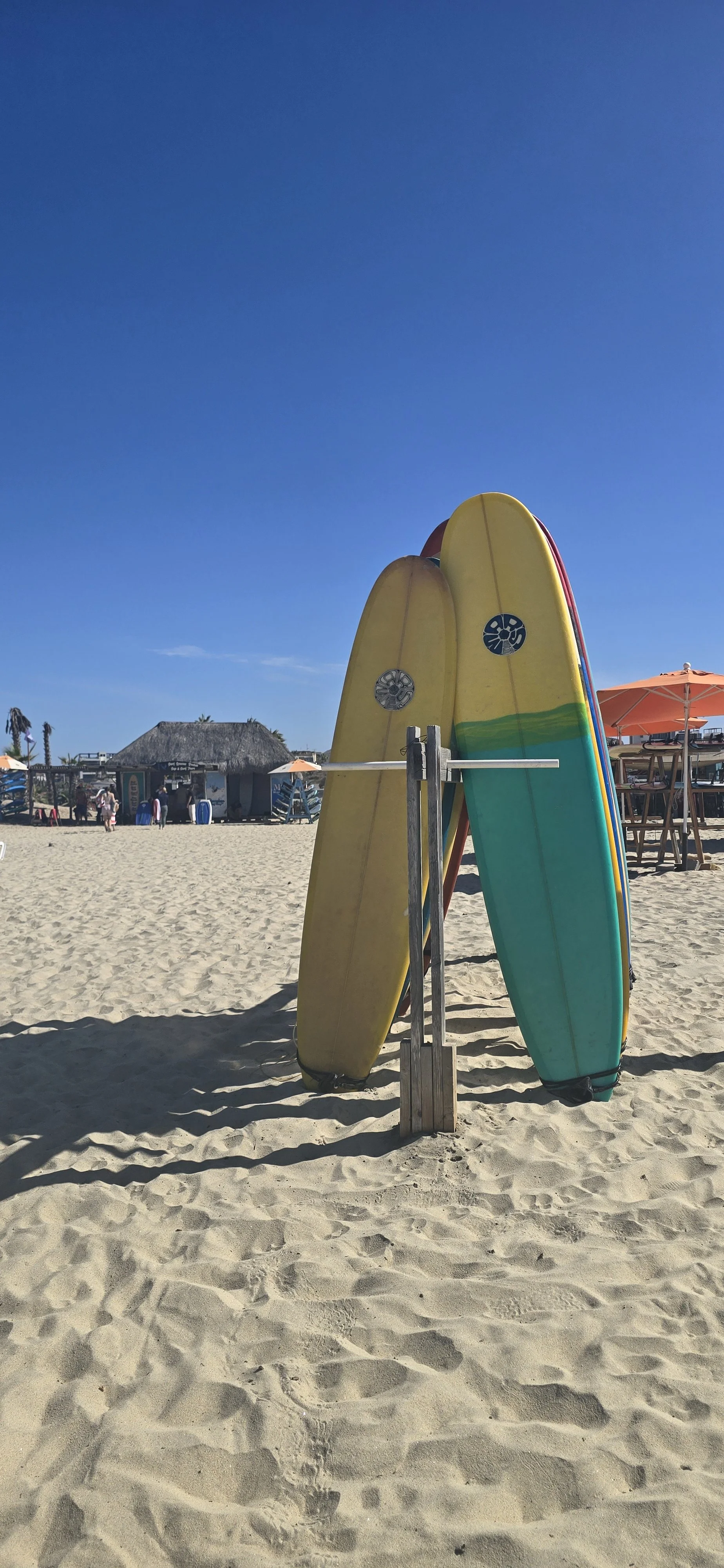 Three surfboards leaning against a wooden stand on a sandy beach with a clear blue sky and beach huts and umbrellas in the background.