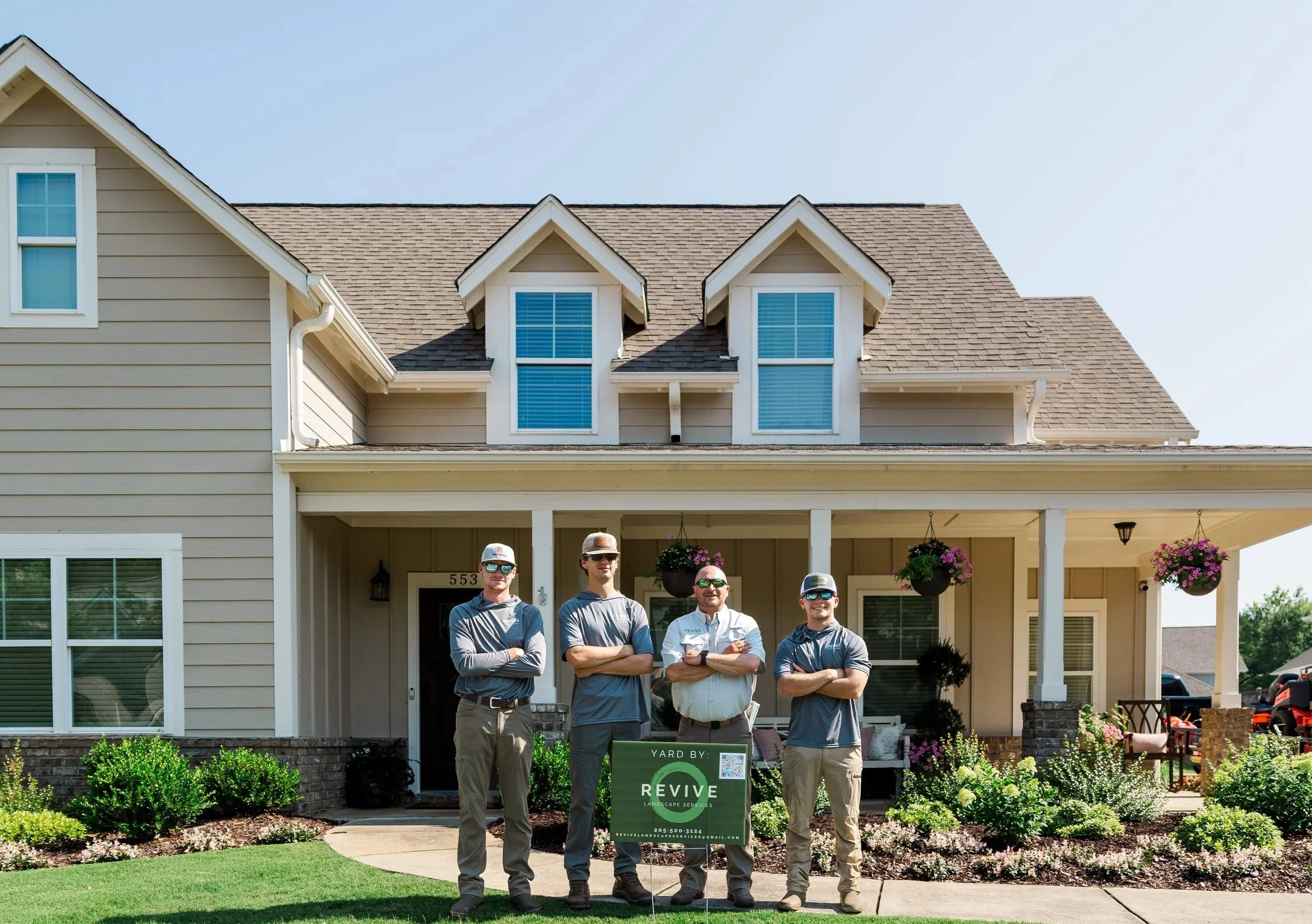 Revive Landscape Helena AL Four people standing in front of a beige house with a gabled roof, holding a sign that says 'Yard by: REVIVE Landscape Services.' The house has large windows, hanging flower baskets, and a neatly maintained garden and lawn.
