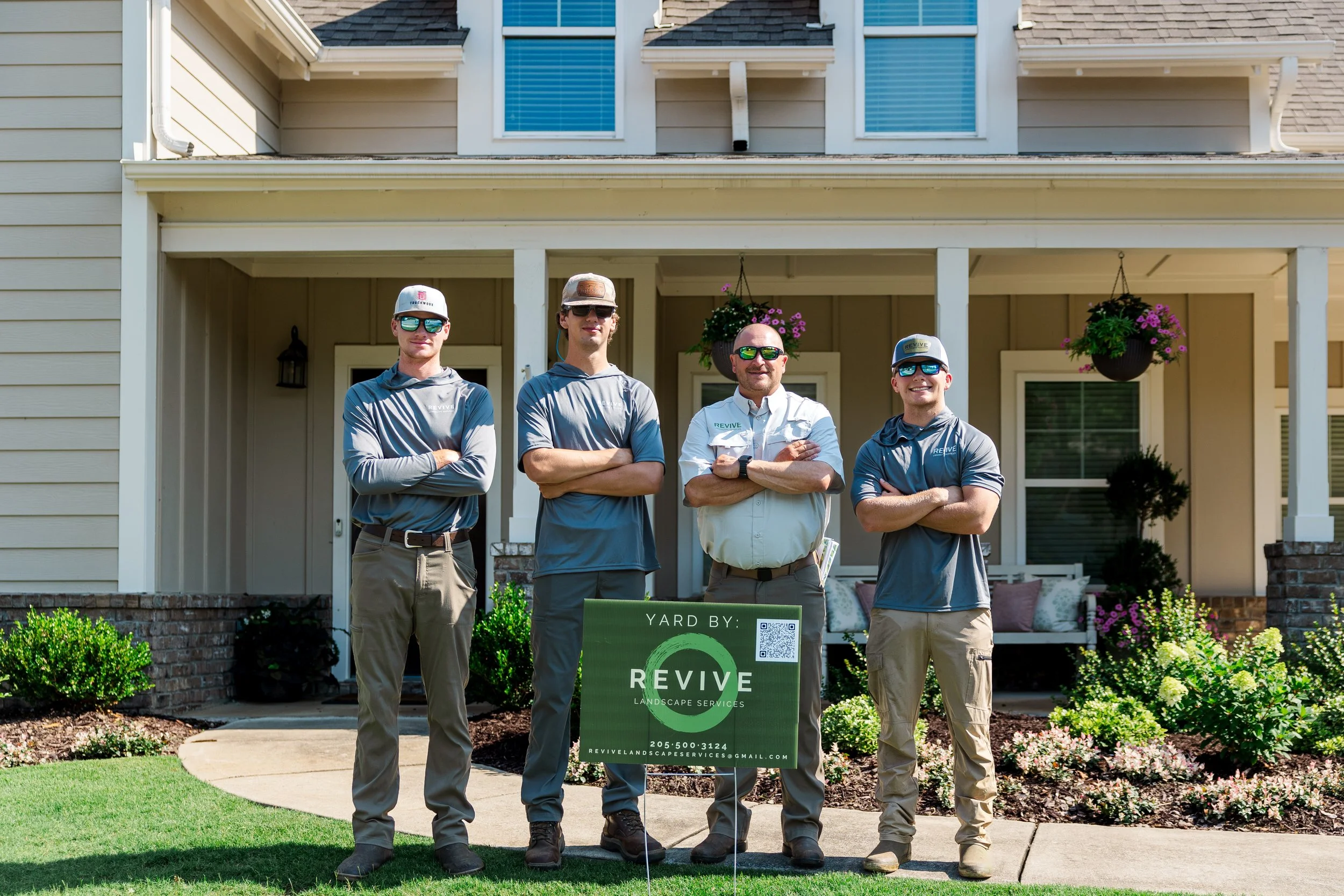 Four men stand in front of a suburban house, posing for a photo. They are wearing golf or workwear style shirts, sunglasses, and hats. There is a green yard sign with the words 'YARD BY: REVIVE LANDSCAPE SERVICES' and contact information. The house has hanging flower baskets, a porch, and a well-maintained lawn.