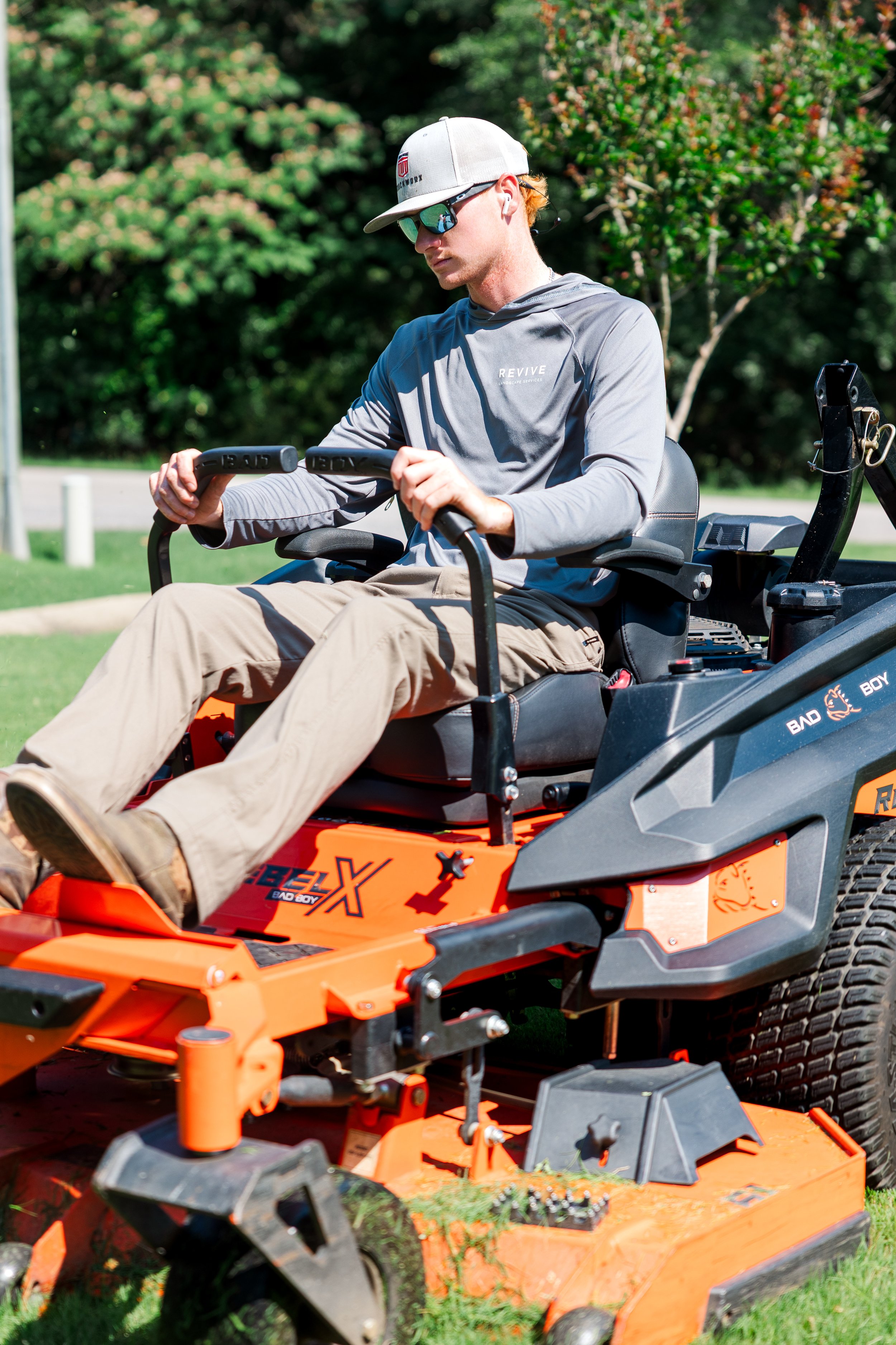 Revive Landscape Helena Alabama A man wearing sunglasses, a cap, and a gray hoodie operating a bright orange and black riding lawn mower on a sunny day with green trees in the background.