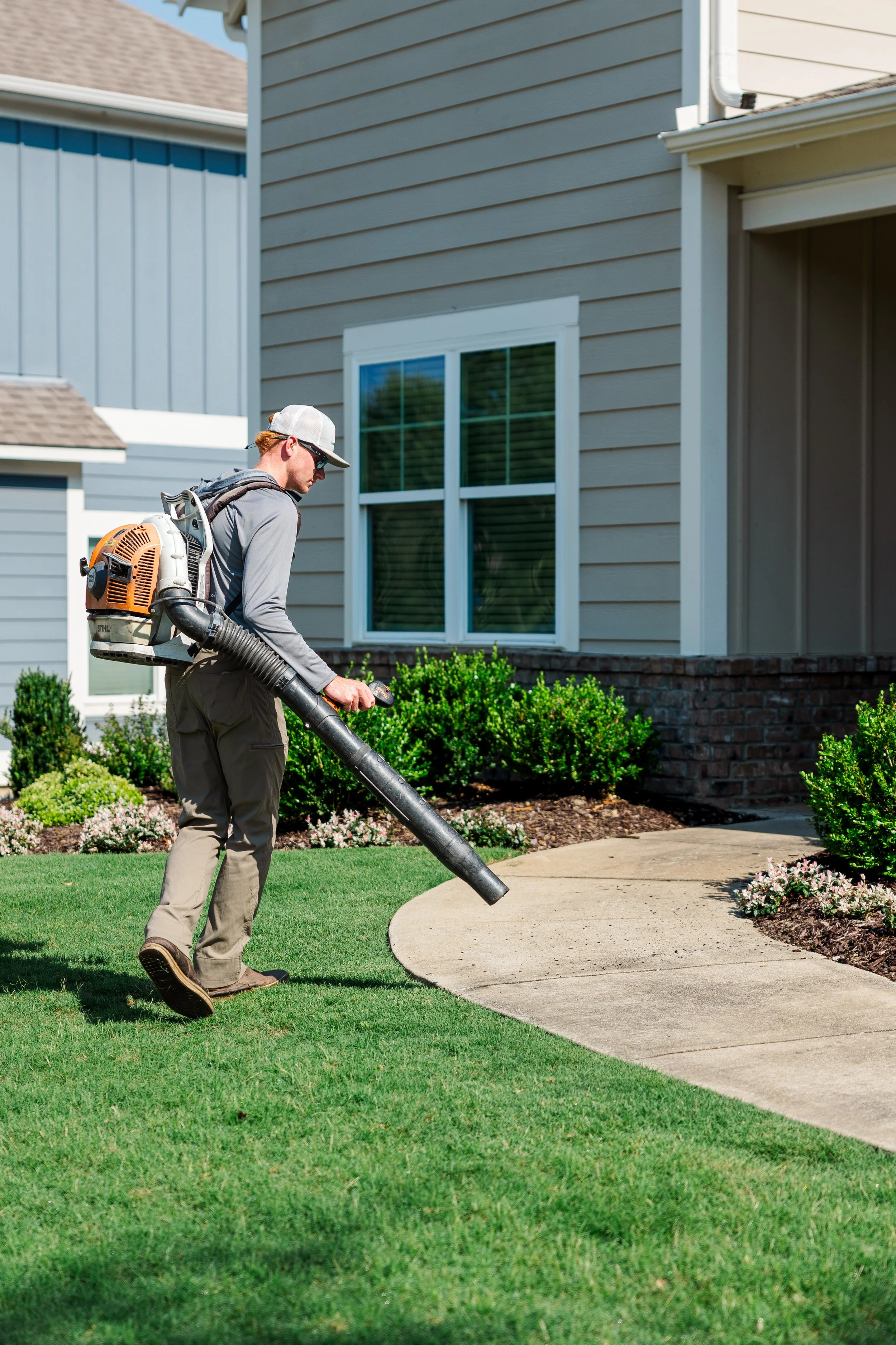 Revive Landscape Helena Alabama A man wearing sunglasses, a cap, and gray work clothes uses a leaf blower to clean a grassy and landscaped front yard of a house with beige siding and a white window.