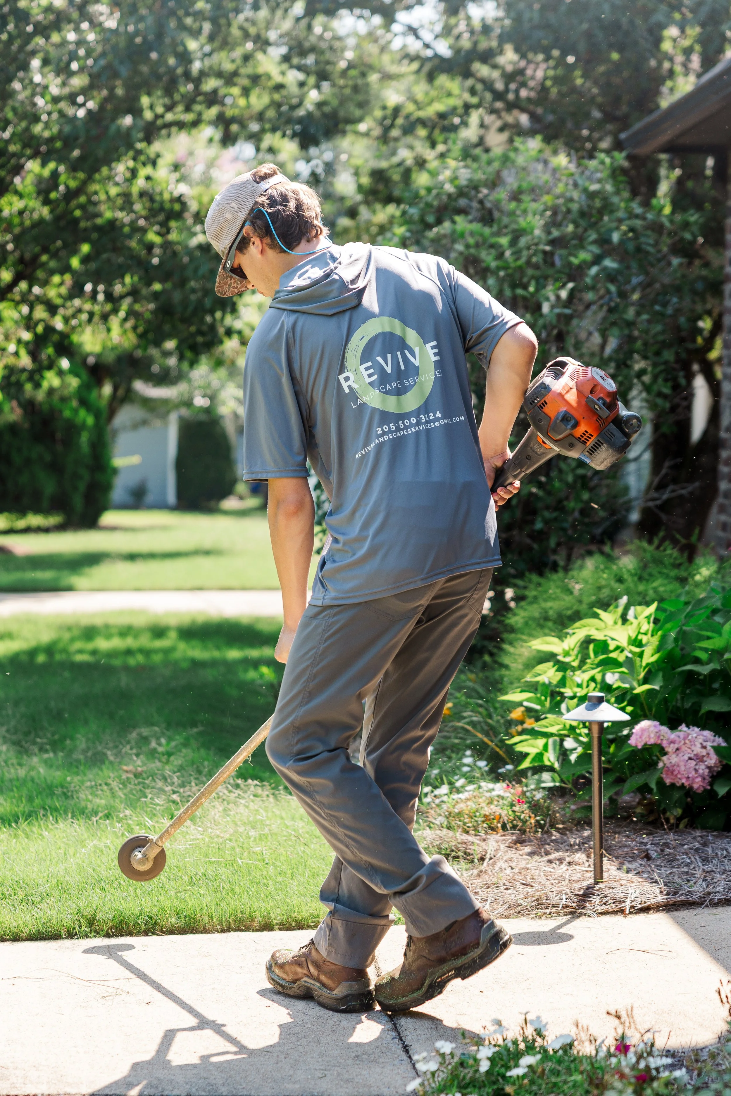 Revive Landscape Helena Alabama A man wearing a gray shirt and brown boots is trimming a garden bed with electric hedge clippers in a sunny backyard with trees and green grass.