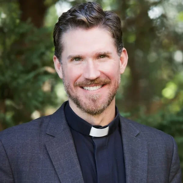 Portrait of a smiling man in clergy attire with a clerical collar, outdoors with greenery in the background.
