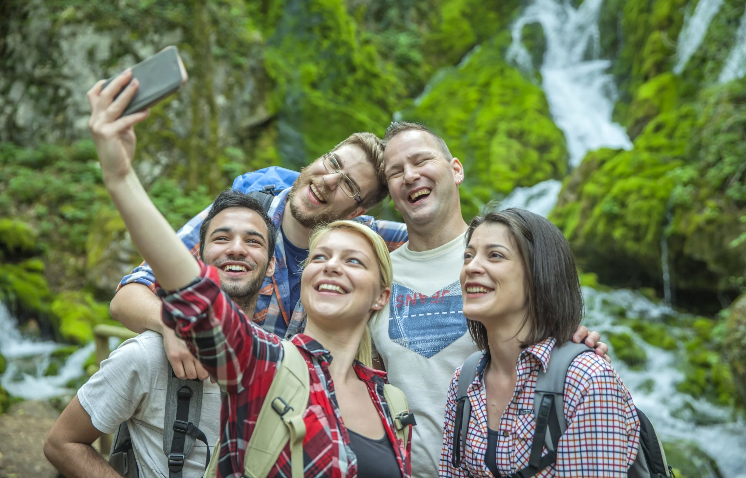 Group of five people taking a selfie in a lush green forest near a waterfall