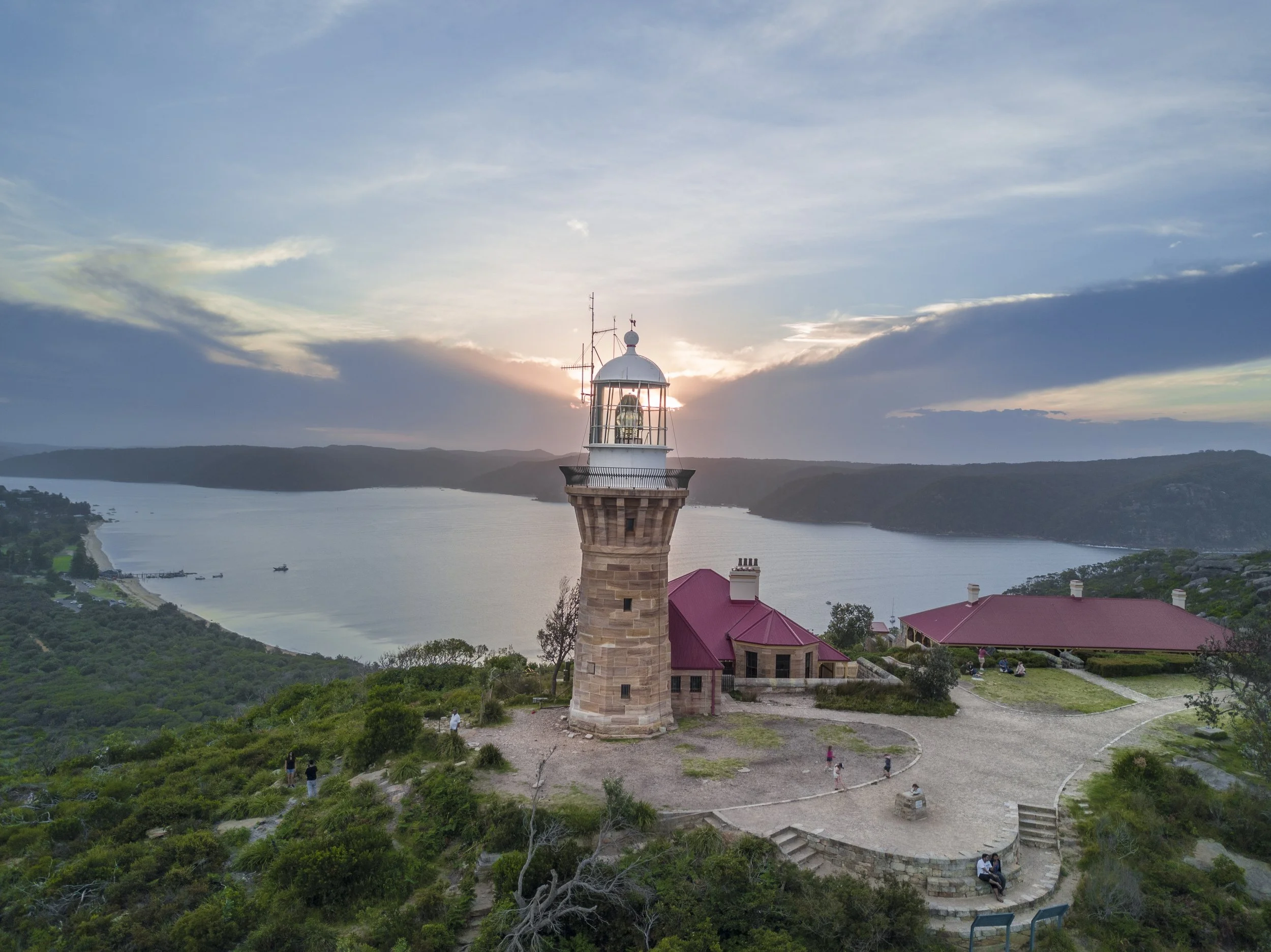 A lighthouse on a hill overlooking a river at sunset with walking paths, trees, and people nearby.