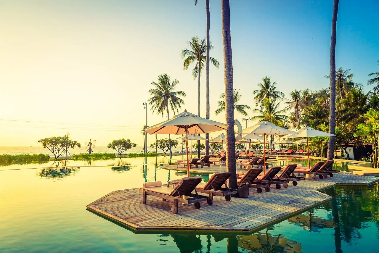 Poolside area with lounge chairs and umbrellas, surrounded by palm trees, overlooking a beach at sunset.