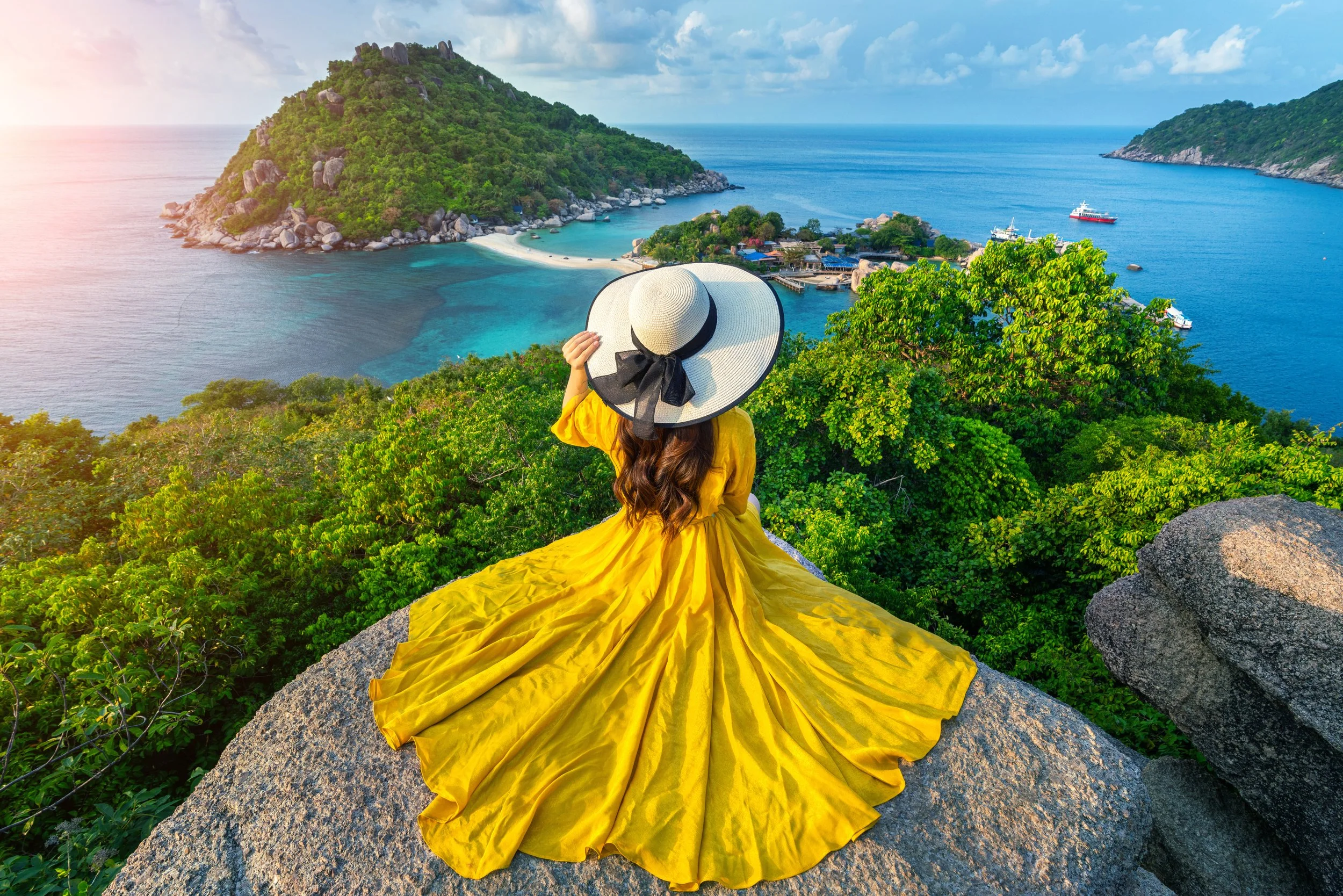 A woman in a yellow dress and a large white sun hat sitting on a rock overlooking a tropical island with a beach, lush greenery, and a blue ocean with boats.