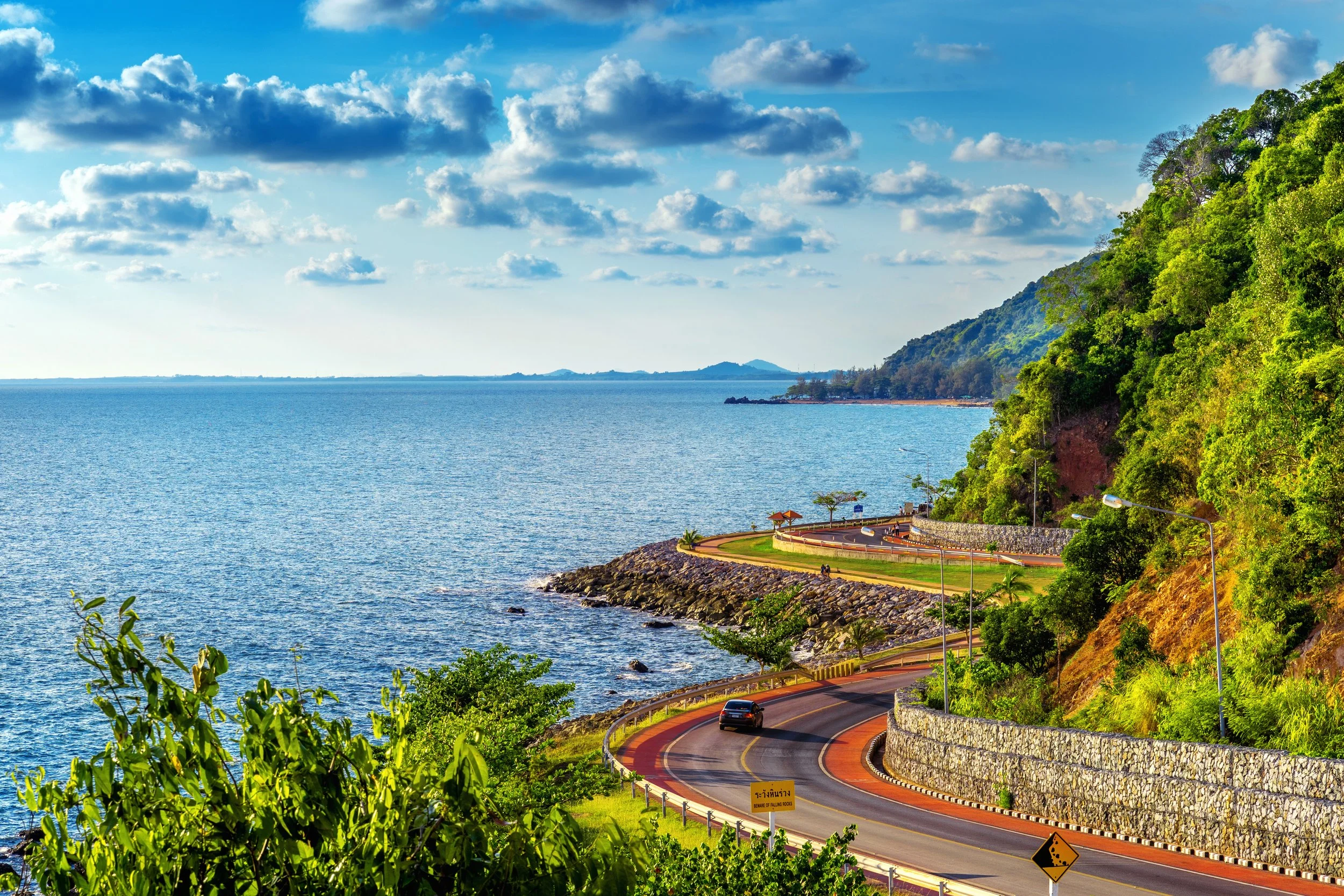 Scenic coastal road winding along green hillside with ocean view, partly cloudy sky, and distant hills.
