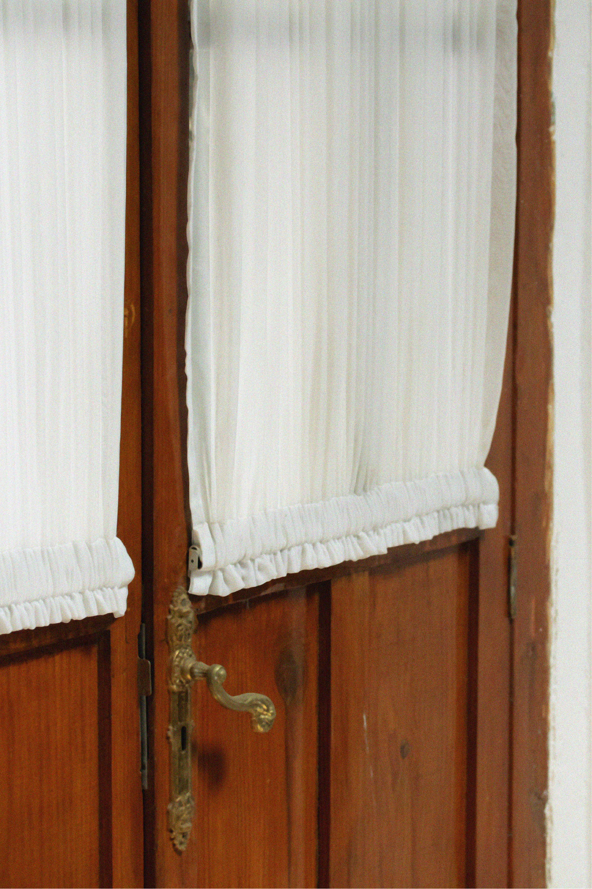 A close-up view of a wooden door with a brass doorknob and a white storm door with a protective screen, featuring a pleated white curtain hanging inside.