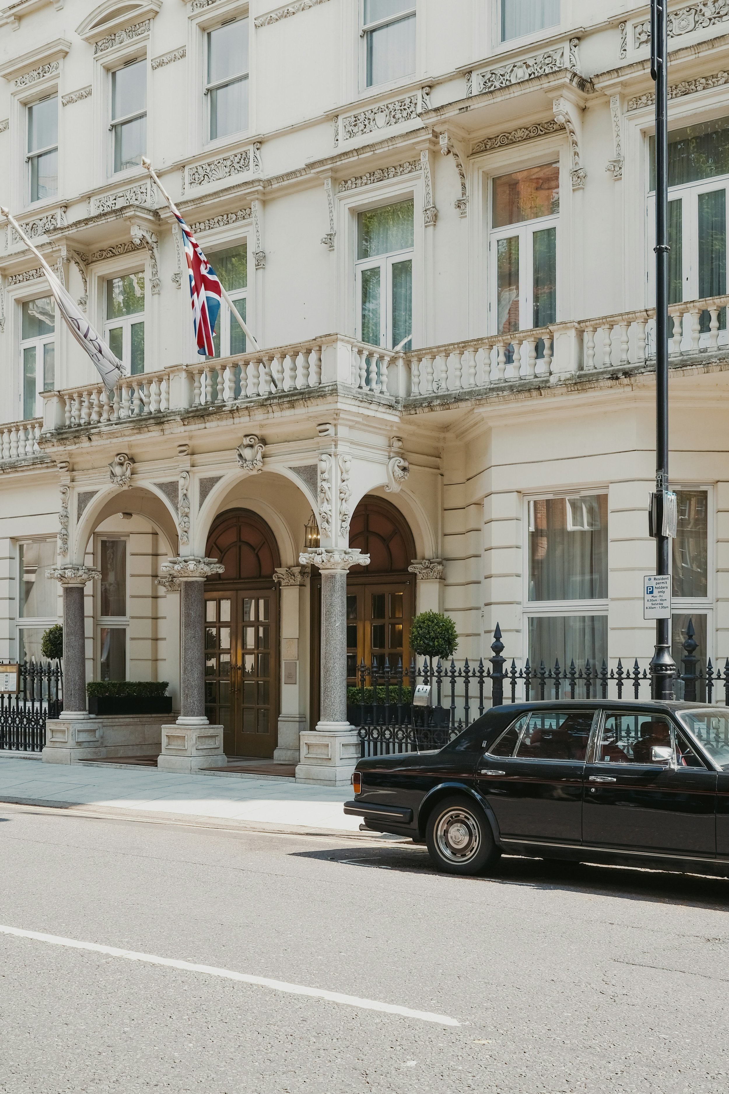 A classic black luxury car parked in front of a white, multi-story building with ornate architectural details, tall windows, and flags on the balcony.