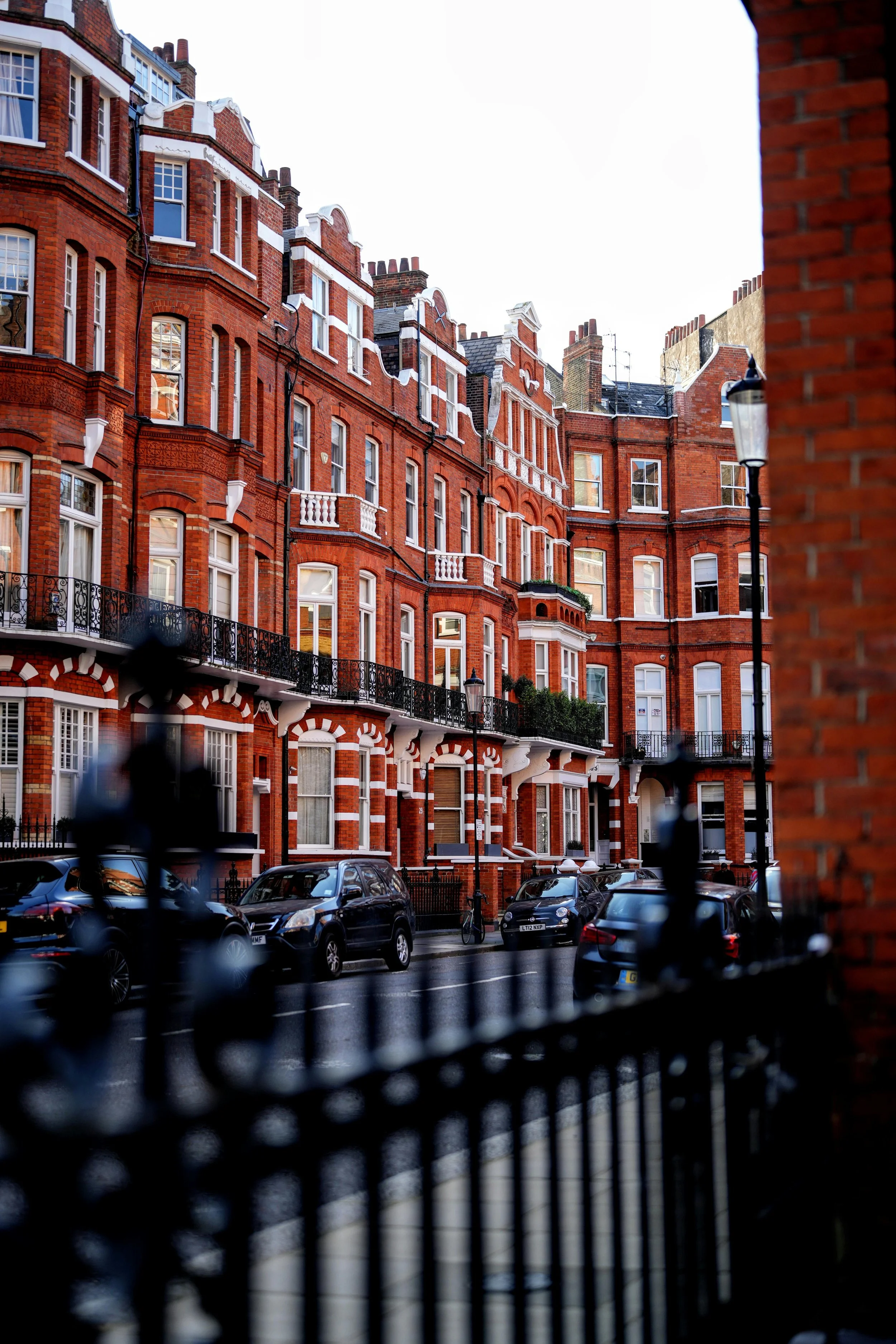 View of red brick residential buildings with black iron railings and parked cars on the street, seen through a black metal fence and a partial brick wall on the right side.