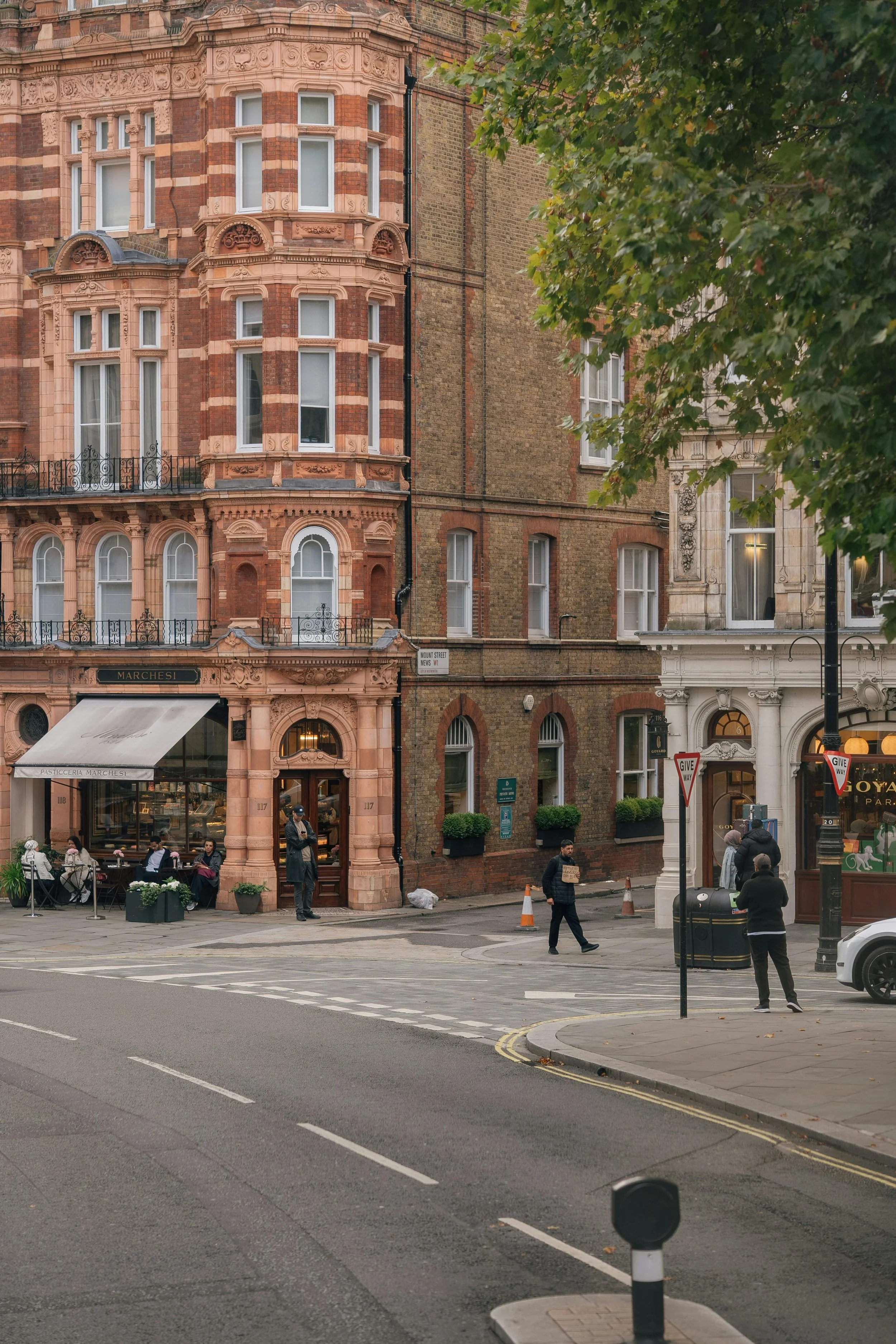 City street view showing a building with ornate architectural details in pinkish terracotta and brick, with a cafe or restaurant with outdoor seating in front, several pedestrians walking, and street signs and traffic cones on the road.