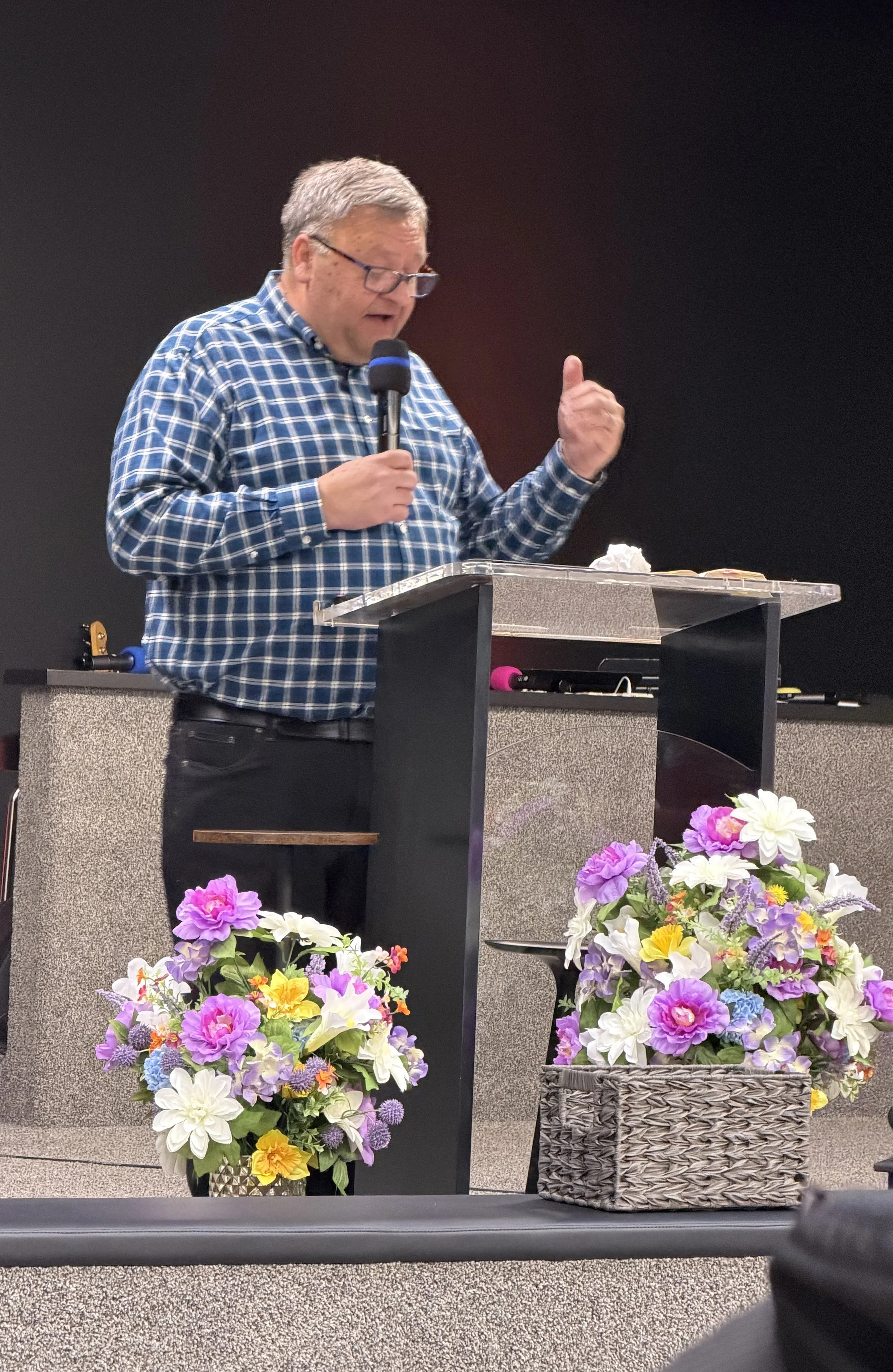 A man in glasses and a blue plaid shirt speaking at a podium with a microphone, with colorful flower arrangements in baskets and pots in front of him, in an indoor setting.