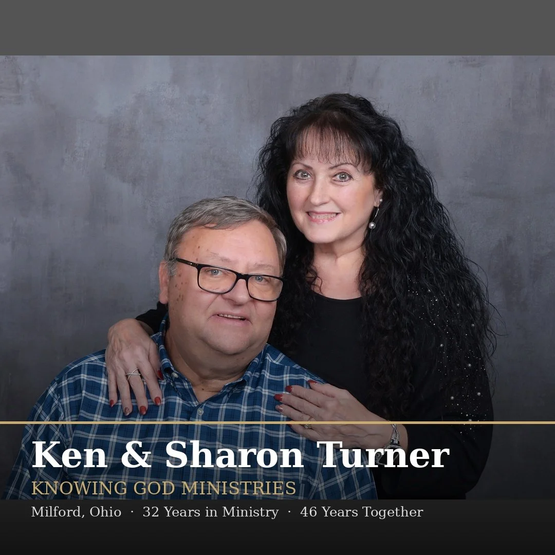 A man with glasses wearing a blue plaid shirt and a woman with black curly hair wearing black attire, smiling, posing in front of a gray backdrop.