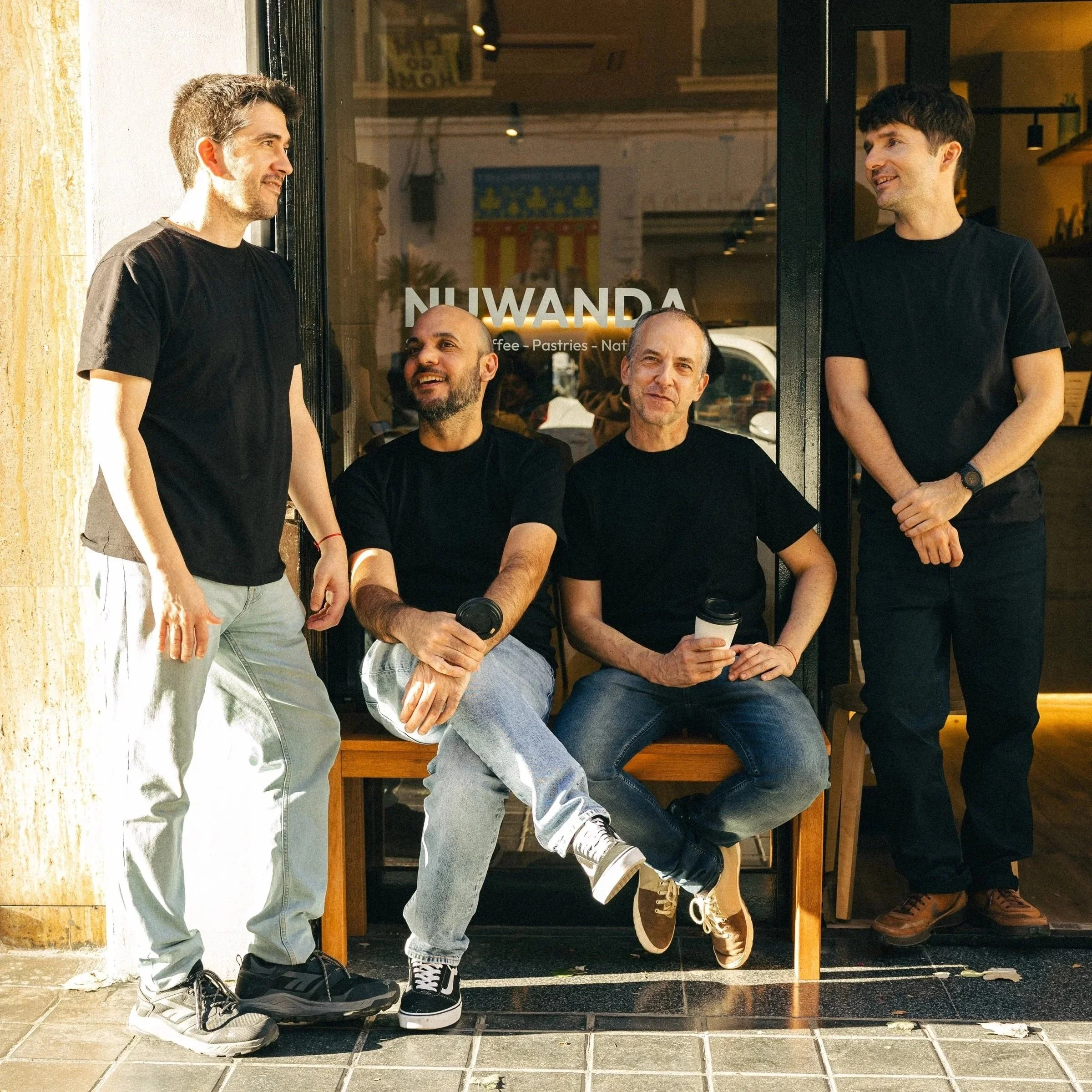 Four men sitting and standing outside a cafe named NIWANDA, with large windows and a door, smiling and engaging in conversations, some holding coffee cups, wearing casual black shirts and jeans.