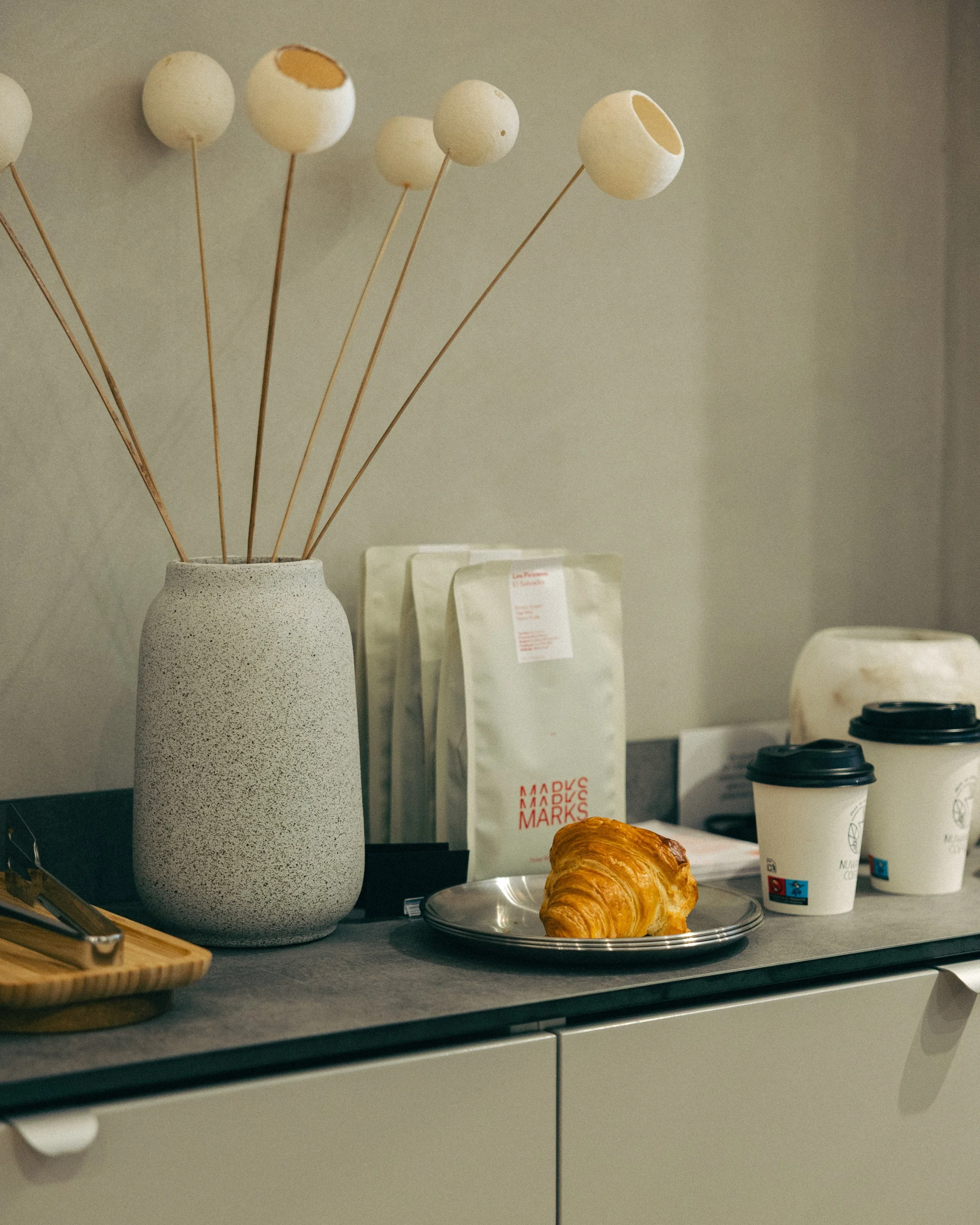 A white textured vase with wooden sticks arranged inside it, some topped with spherical objects. On the counter, a croissant on a silver tray, a paper bag with red and white lettering, and two cups of coffee in paper cups with black plastic lids. In the background, there are menu cards.