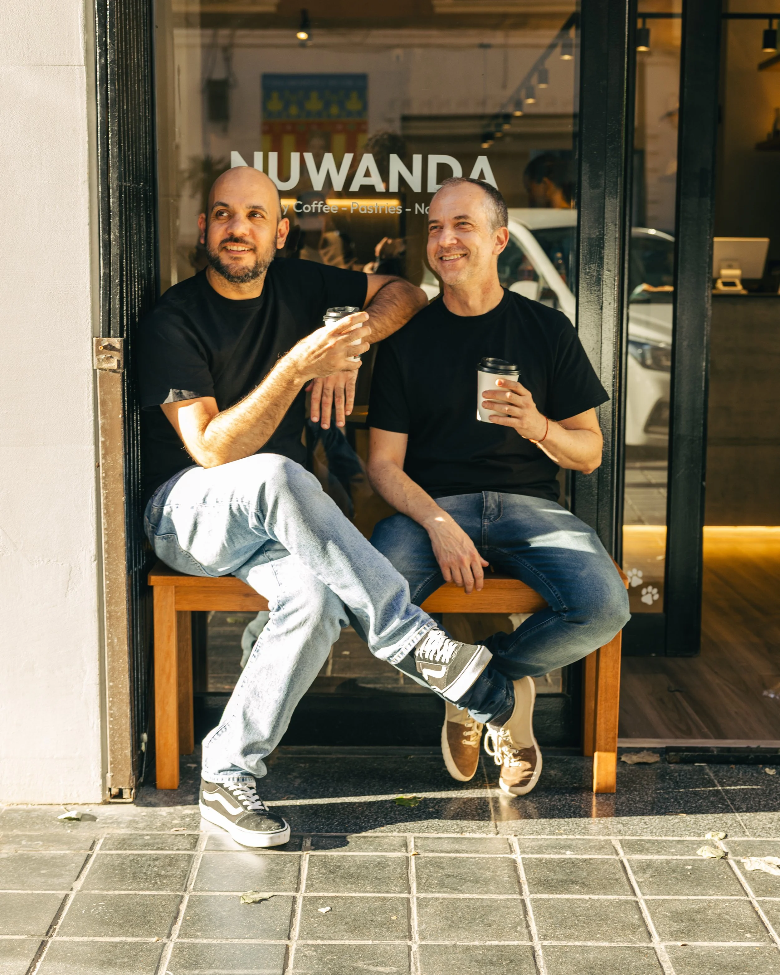 Two men sitting at a cafe window, holding coffee cups, smiling, and wearing black t-shirts and jeans. One man has his arm around the other's shoulder. The window has the word 'NUWANDA' and mentions coffee and pastries.