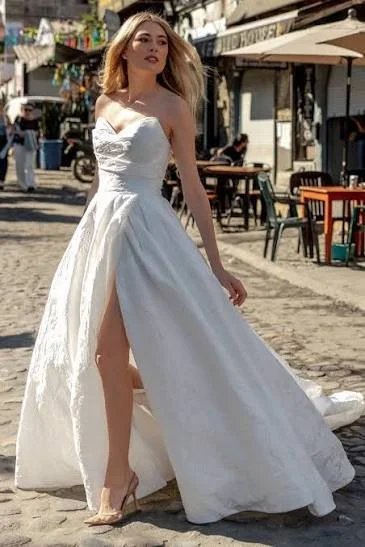Woman in a white wedding dress with an open slit, standing on a cobblestone street with outdoor seating and shops in the background.