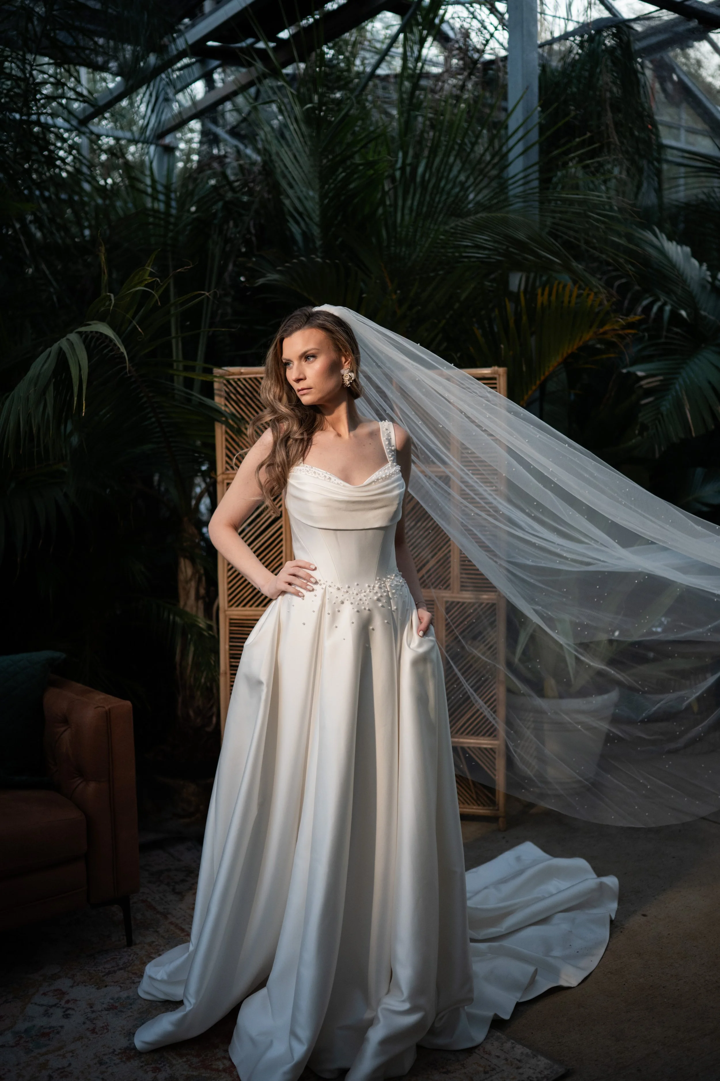 A woman in an elegant white wedding dress with a long train and veil, standing indoors among lush green plants and wooden furniture with a decorative screen in the background.