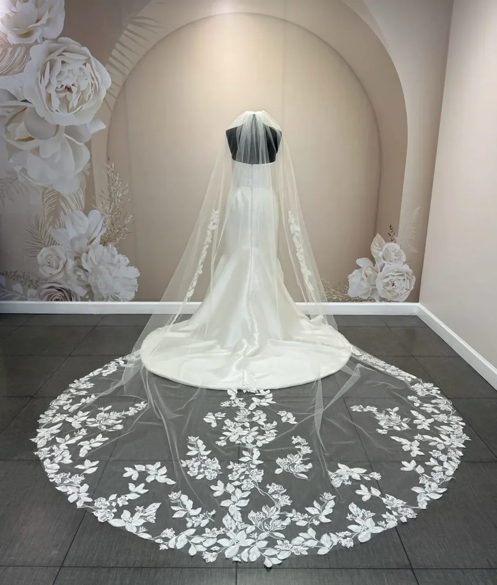 Display of a wedding dress on a mannequin with a long, lace-embellished veil and train, set against a decorated wall with large white floral paper decorations.