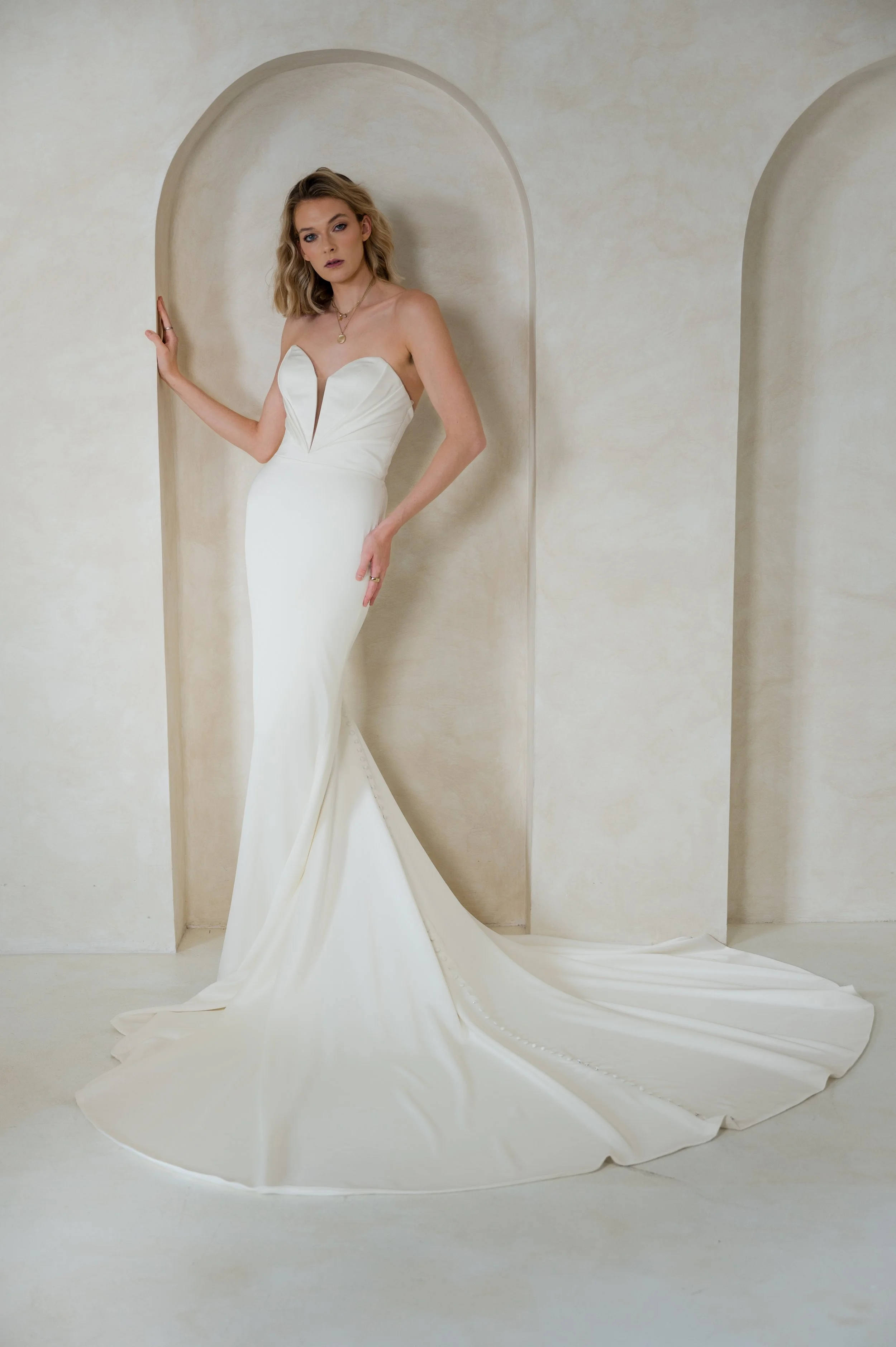 A woman in a white strapless wedding gown with a sweetheart neckline, standing in a minimalist cream-colored room with arched wall niches, and a long train flowing on the floor.