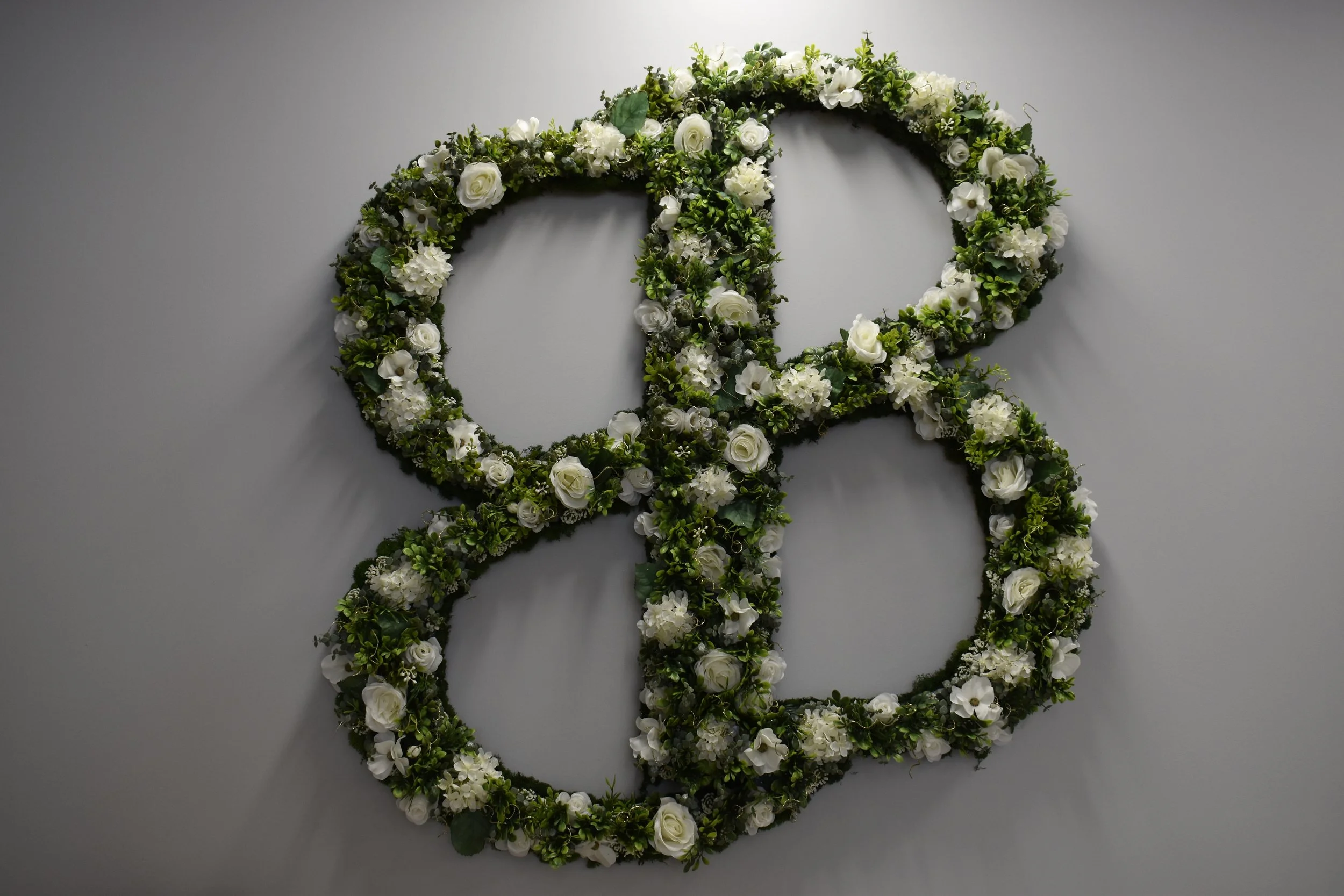 A floral gender reveal wreath made of white and green flowers hanging on a plain gray wall.