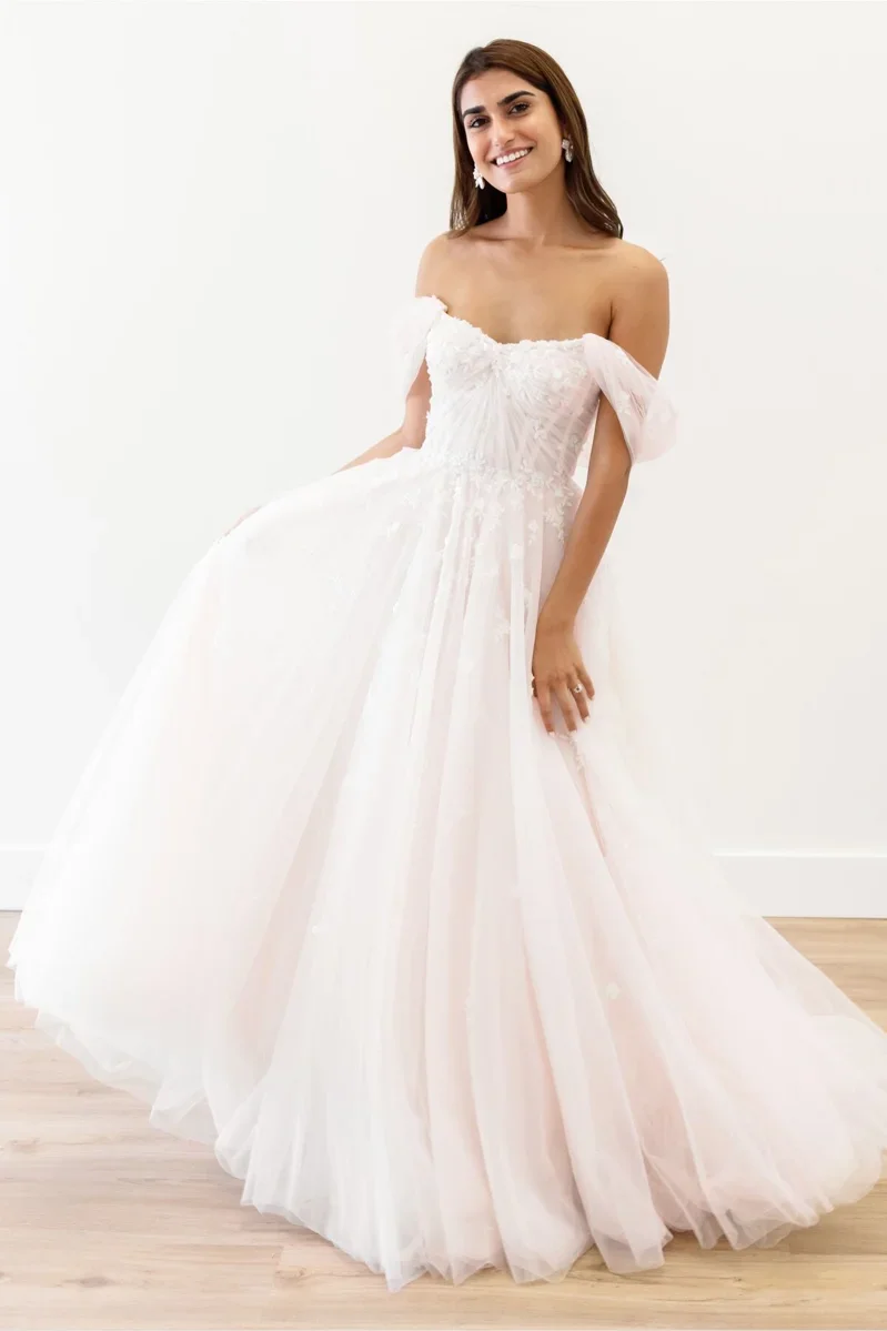 A woman wearing a strapless white wedding gown with lace details, puffed sleeves, and a full tulle skirt, standing against a white wall with wooden flooring, smiling at the camera.