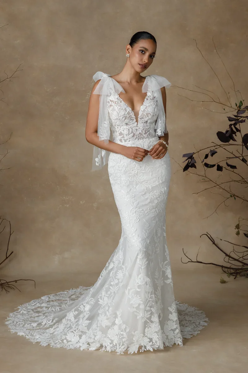 Woman in a white lace wedding dress with a fitted silhouette, sitting against a beige background with minimal plants.