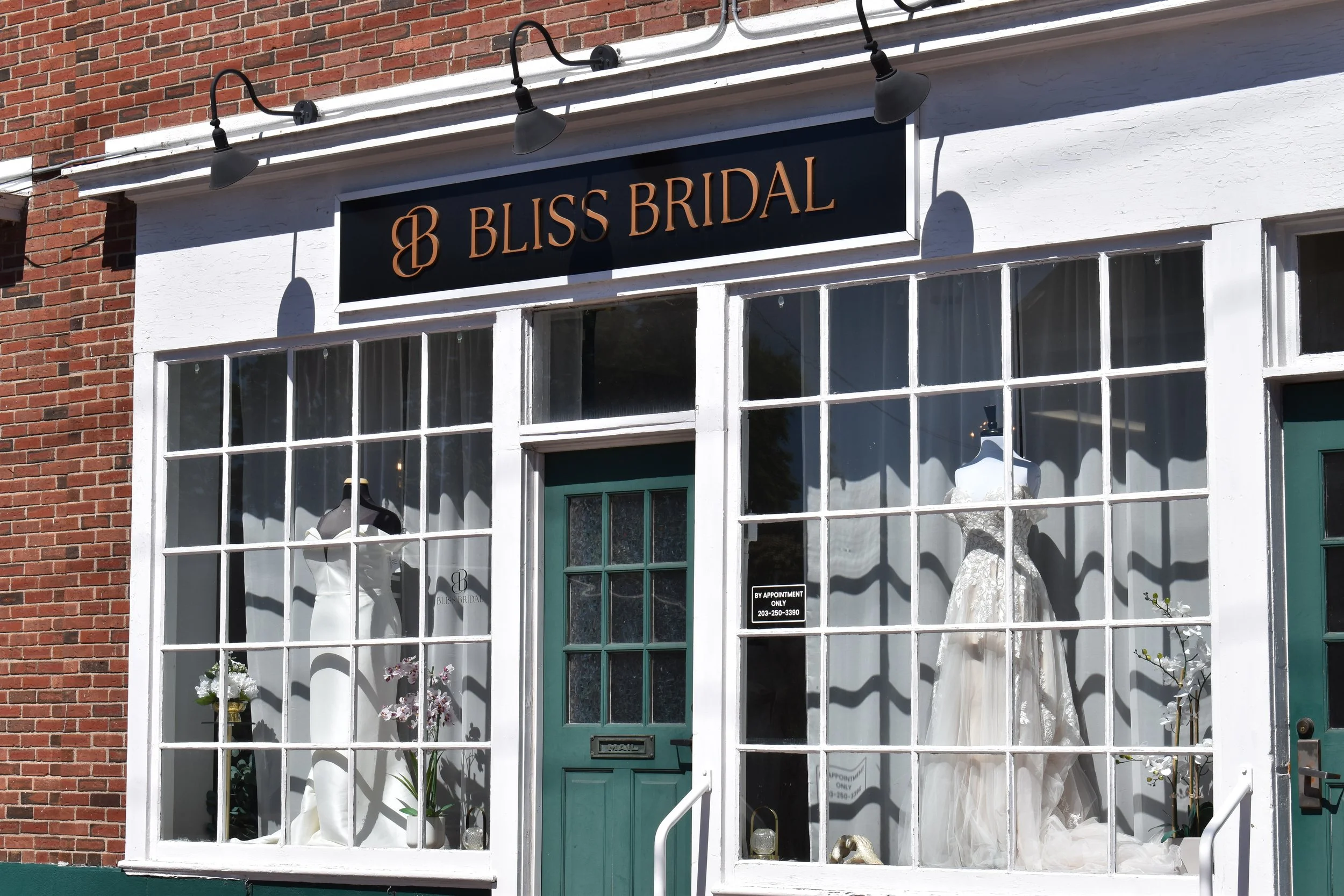 View of a bridal boutique storefront with a black sign reading 'Bliss Bridal' in gold lettering. Display window showcases wedding dresses, mannequins, and flowers inside. The building has a brick wall with a white trim around the window.