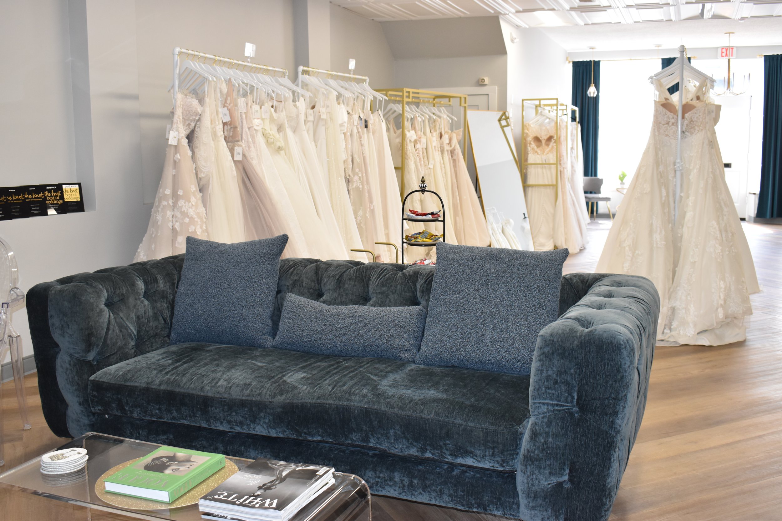 A boutique display of wedding dresses on racks in a bright, modern store. The dresses are in light colors, mostly white or ivory, with lace and floral details. There is a seating area with a plush dark gray sofa and decorative pillows in front of the dresses. A clear coffee table holds magazines and books.