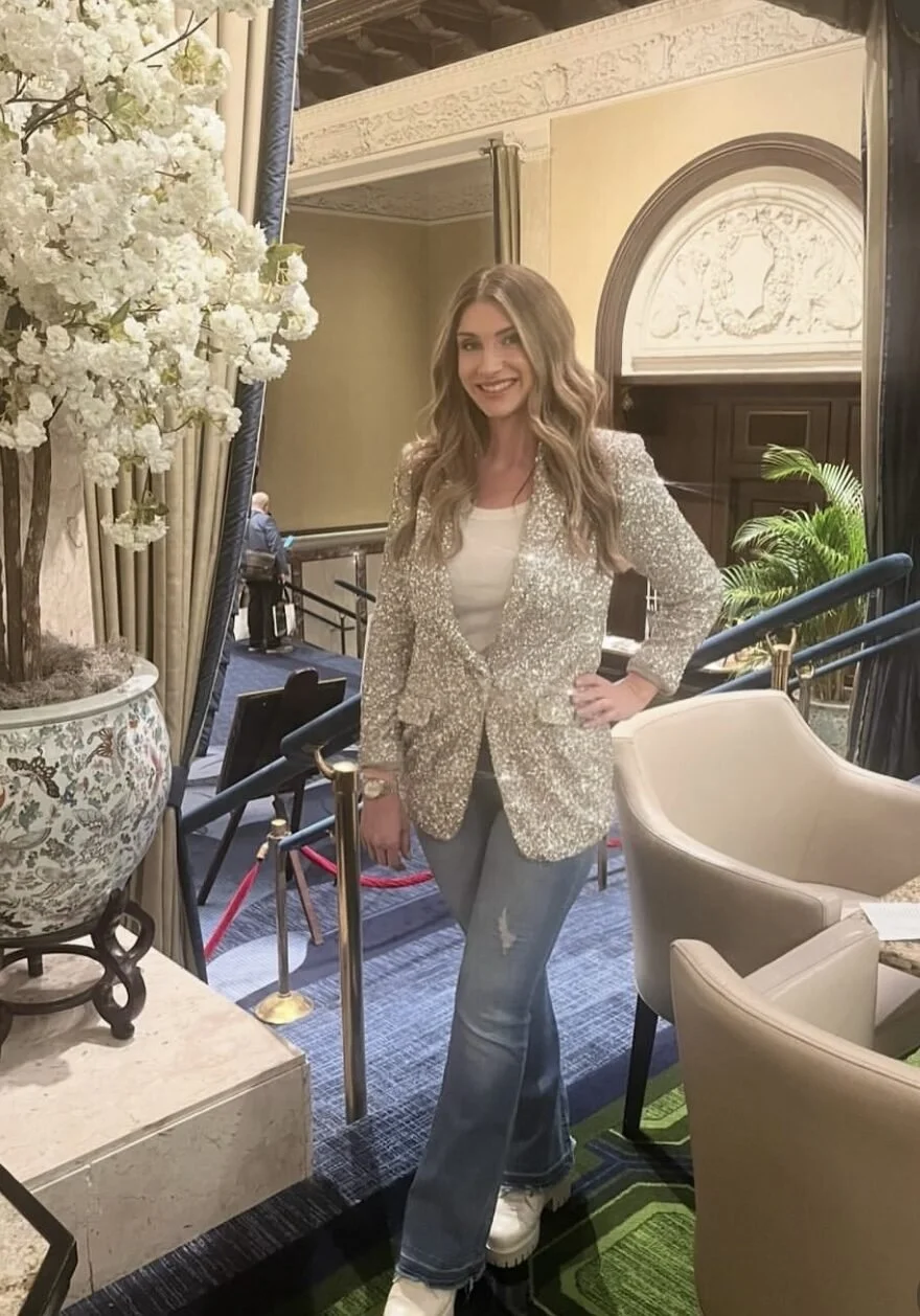 A woman with long wavy hair smiling, wearing a glittery blazer, white top, ripped jeans, and white shoes, standing in a luxurious indoor setting with elegant decor, large floral arrangement, and patterned carpet.