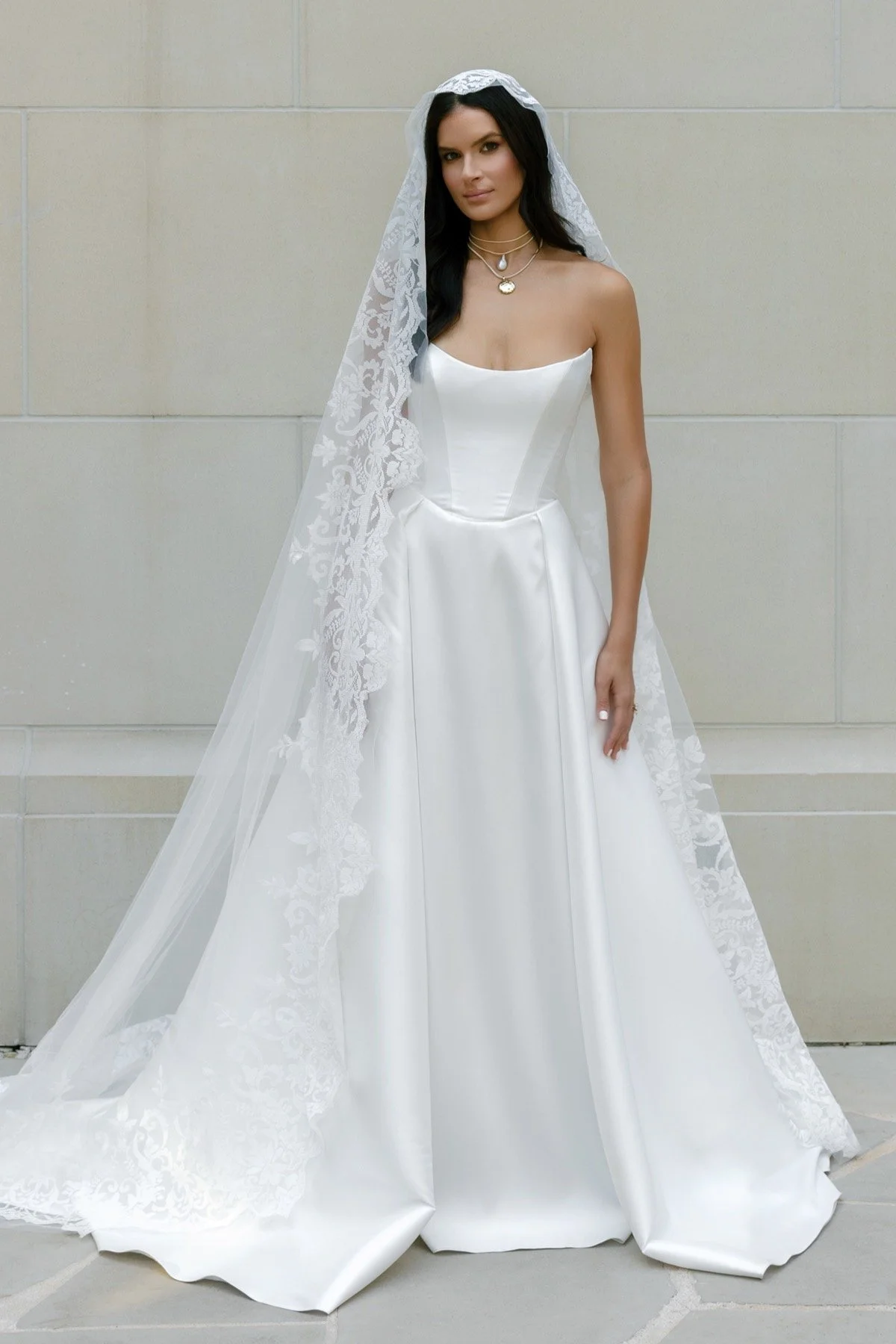 A woman in a white satin wedding dress with lace veil standing against a beige wall.