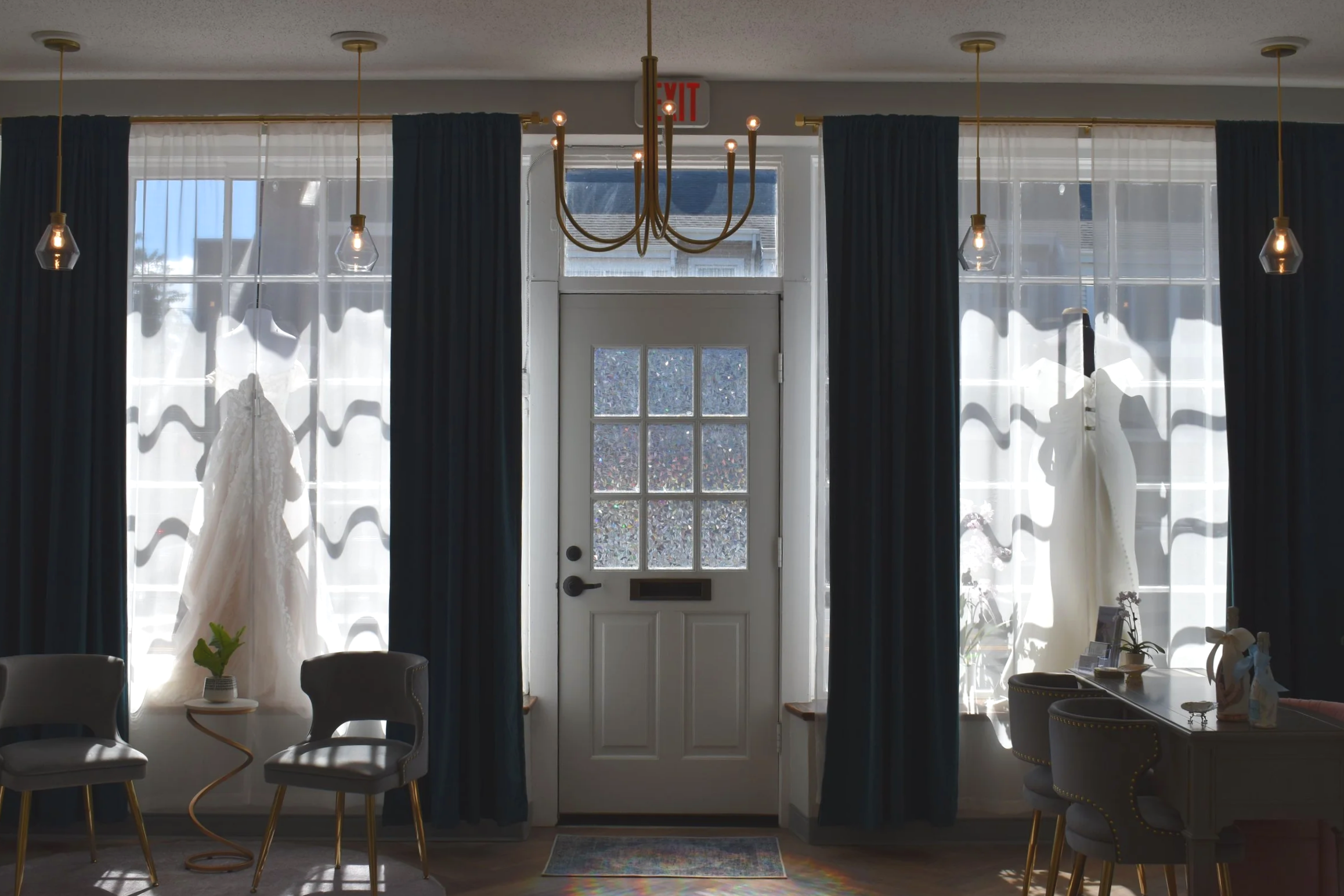 Interior view of a living space with dark curtains, white sheer curtains, and a front door with glass panels. Decorations include a chandelier, pendant lights, a wedding dress hanging on the left, chairs, and small tables with plants and miscellaneous items.