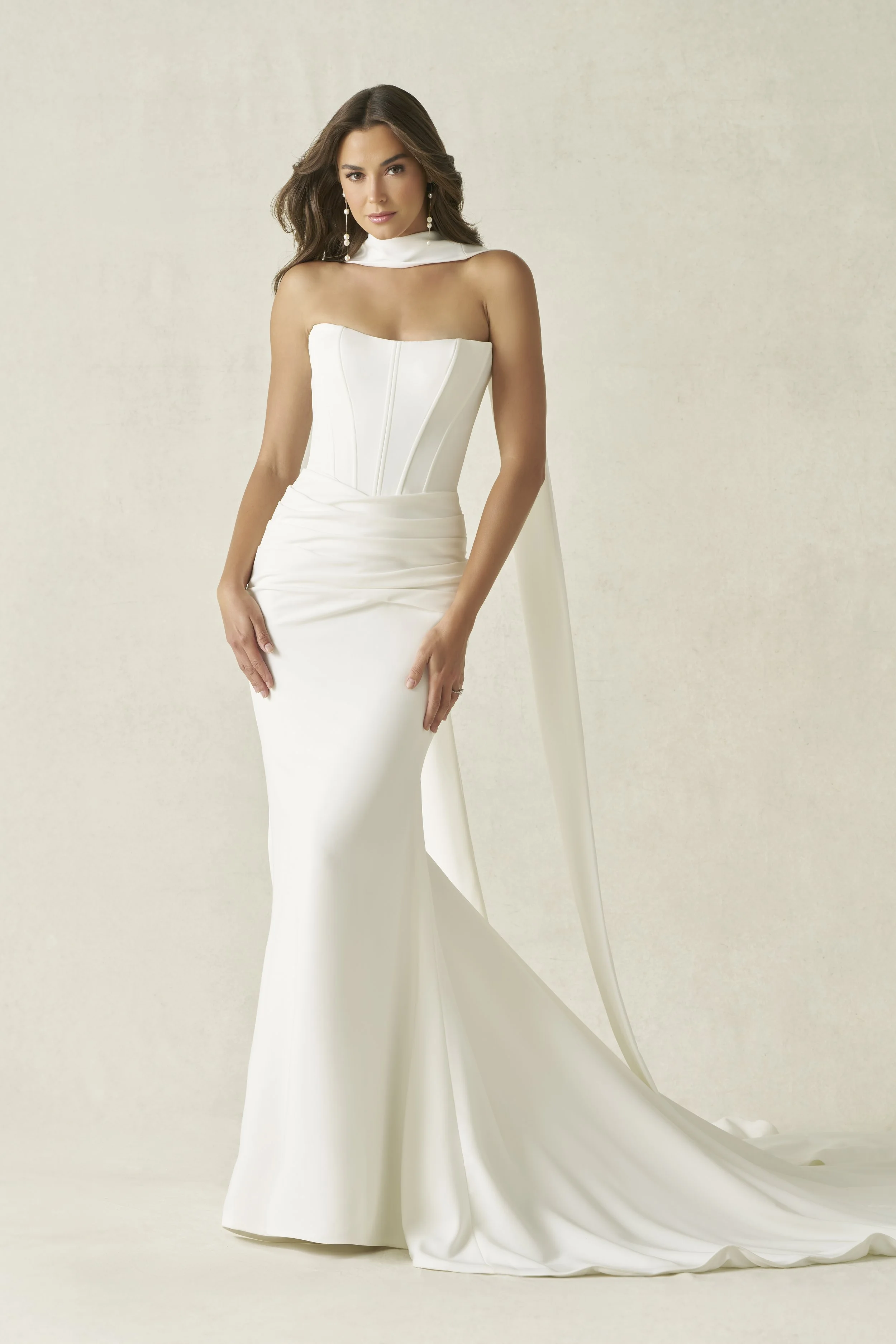 Woman in a strapless white wedding gown with a high slit, standing against a neutral background, with long hair and earrings.