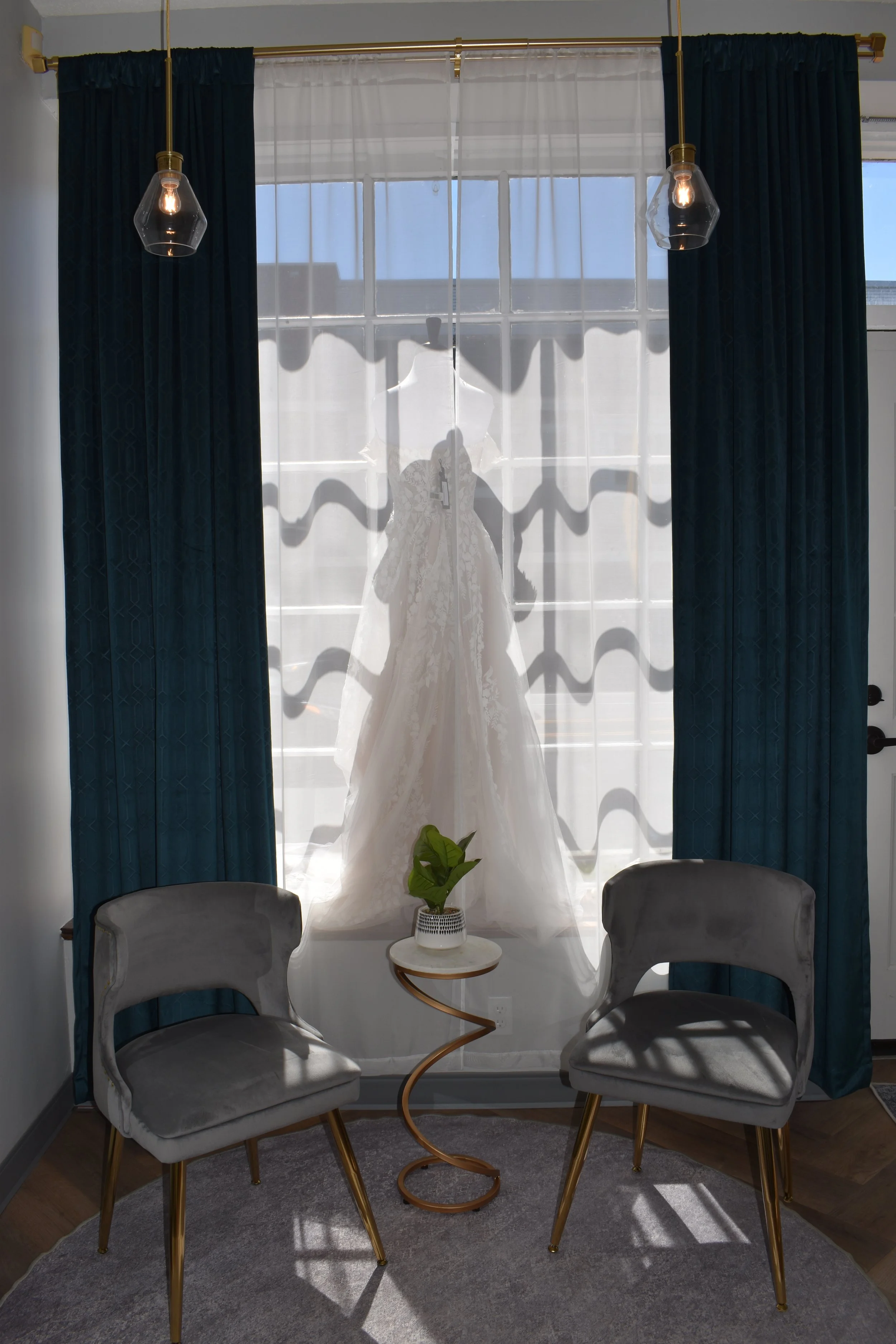 indow with dark blue curtains, a white sheer curtain, and a wedding dress hanging in front of the window. There are two gray chairs with gold legs and a small white table with a green plant in a white pot. The room has a gray carpet and some sunlight coming through the window.