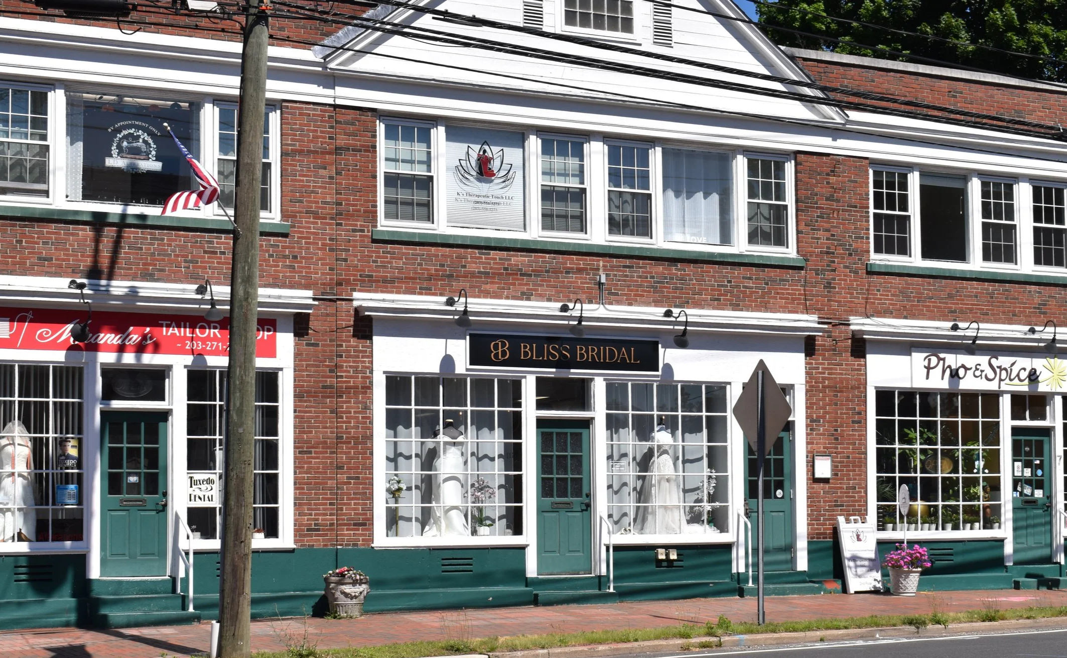 Street view of a brick building with white-framed windows and storefronts. Visible signs include 'Bliss Bridal' and part of a wedding dress display in the window. An American flag is mounted on a pole in front of the building, casting a shadow. There is a wooden utility pole and electrical wires in the foreground, along with potted plants and sidewalk decorations.