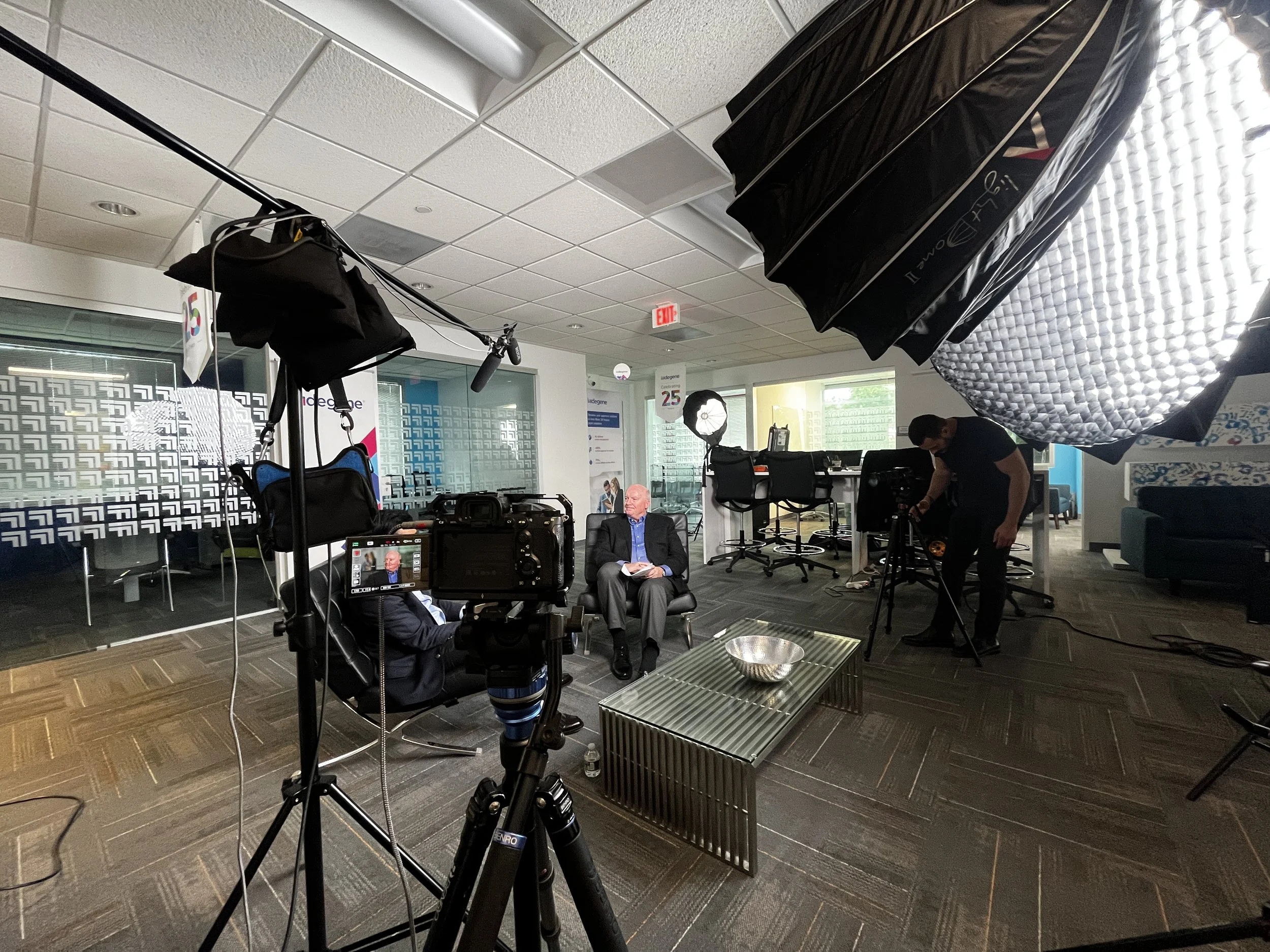 A TV studio or media interview room set up for filming. There is a man in a suit sitting in a black chair, holding papers, with a camera and lighting equipment directed towards him. The room has chairs, banners, and a window with blinds.
