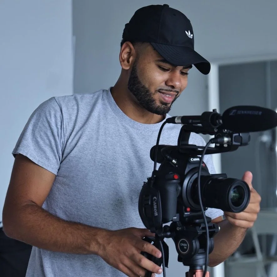 Young man wearing a black cap and gray t-shirt adjusting a professional camera with a microphone attached.