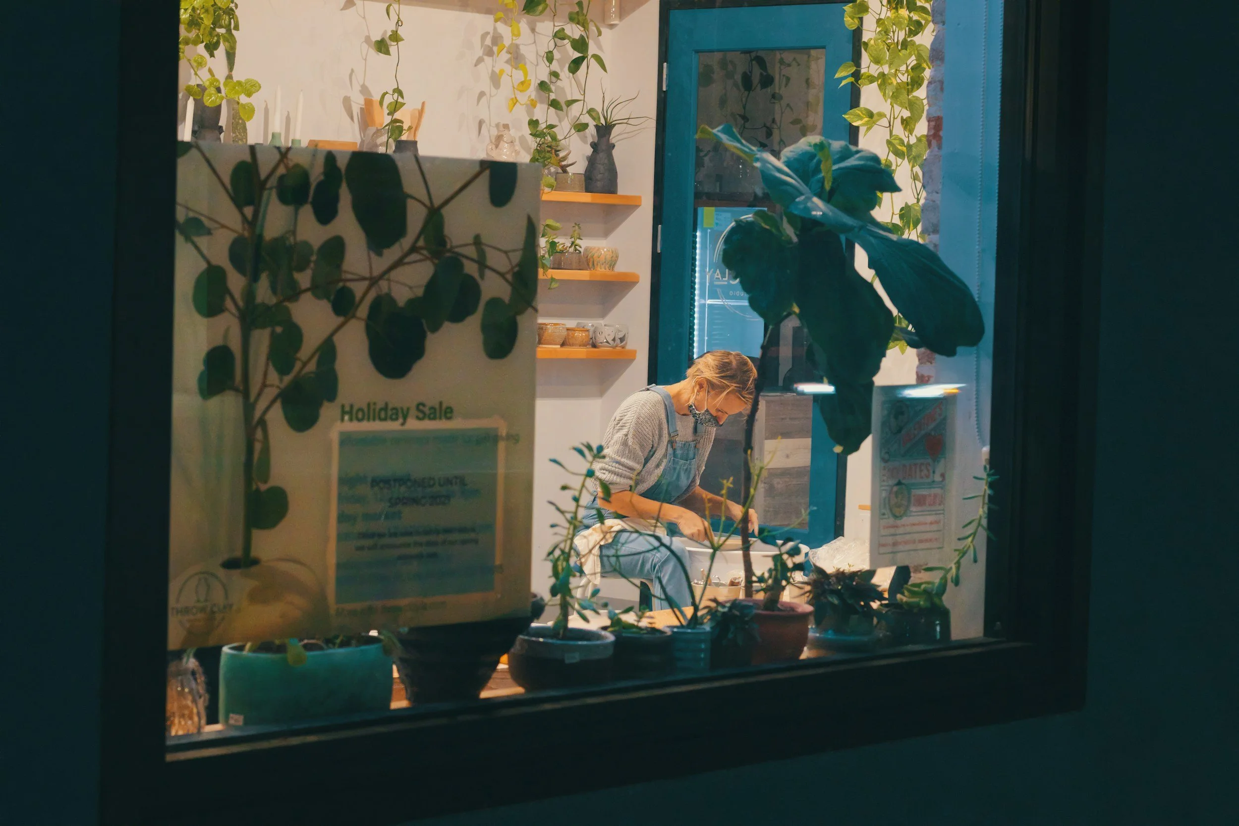 View through a window of a person working inside a shop, surrounded by plants and shelves with decorative items.