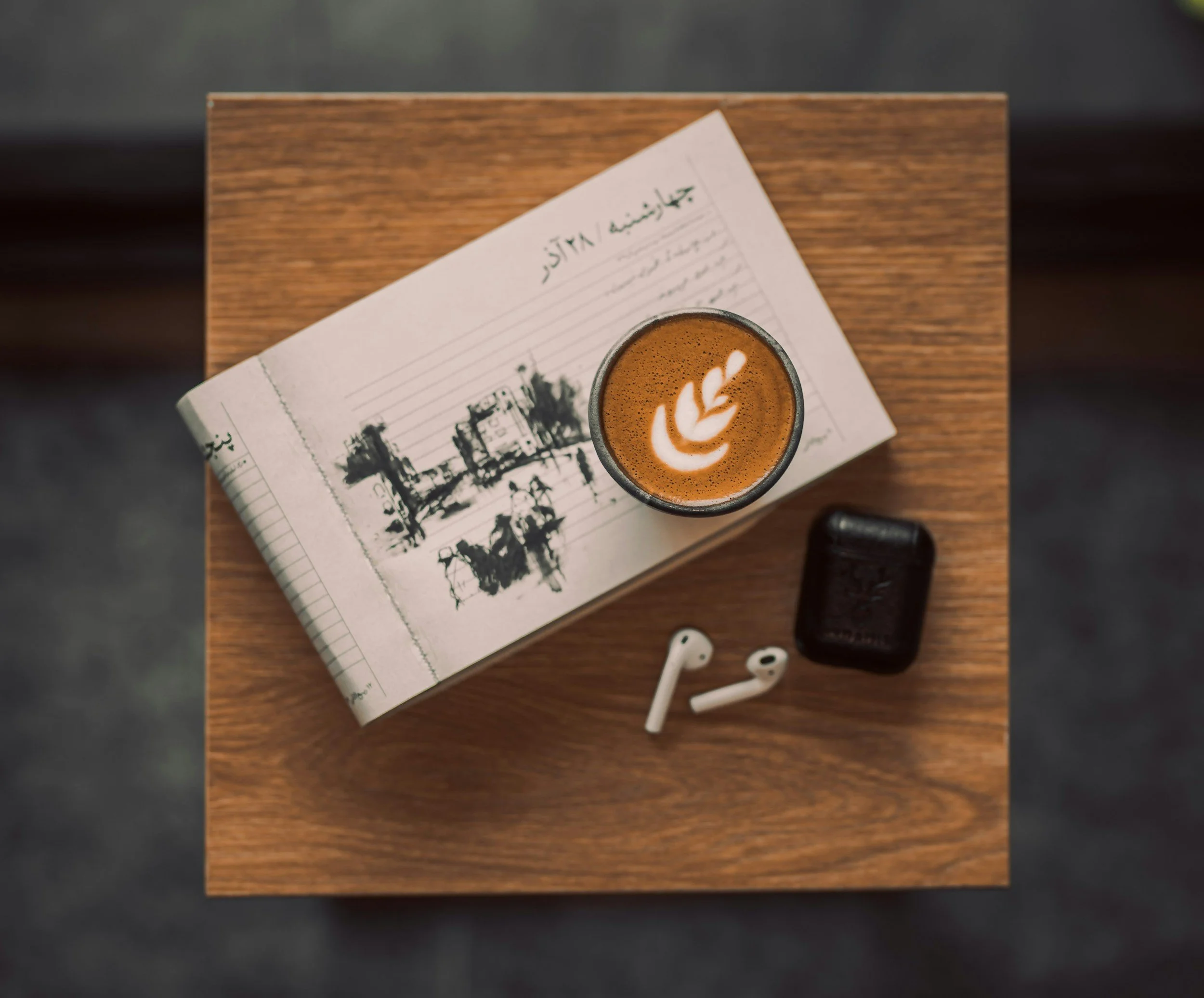 Top view of a wooden table with a newspaper, a cup of coffee with latte art, and a pair of wireless earbuds.