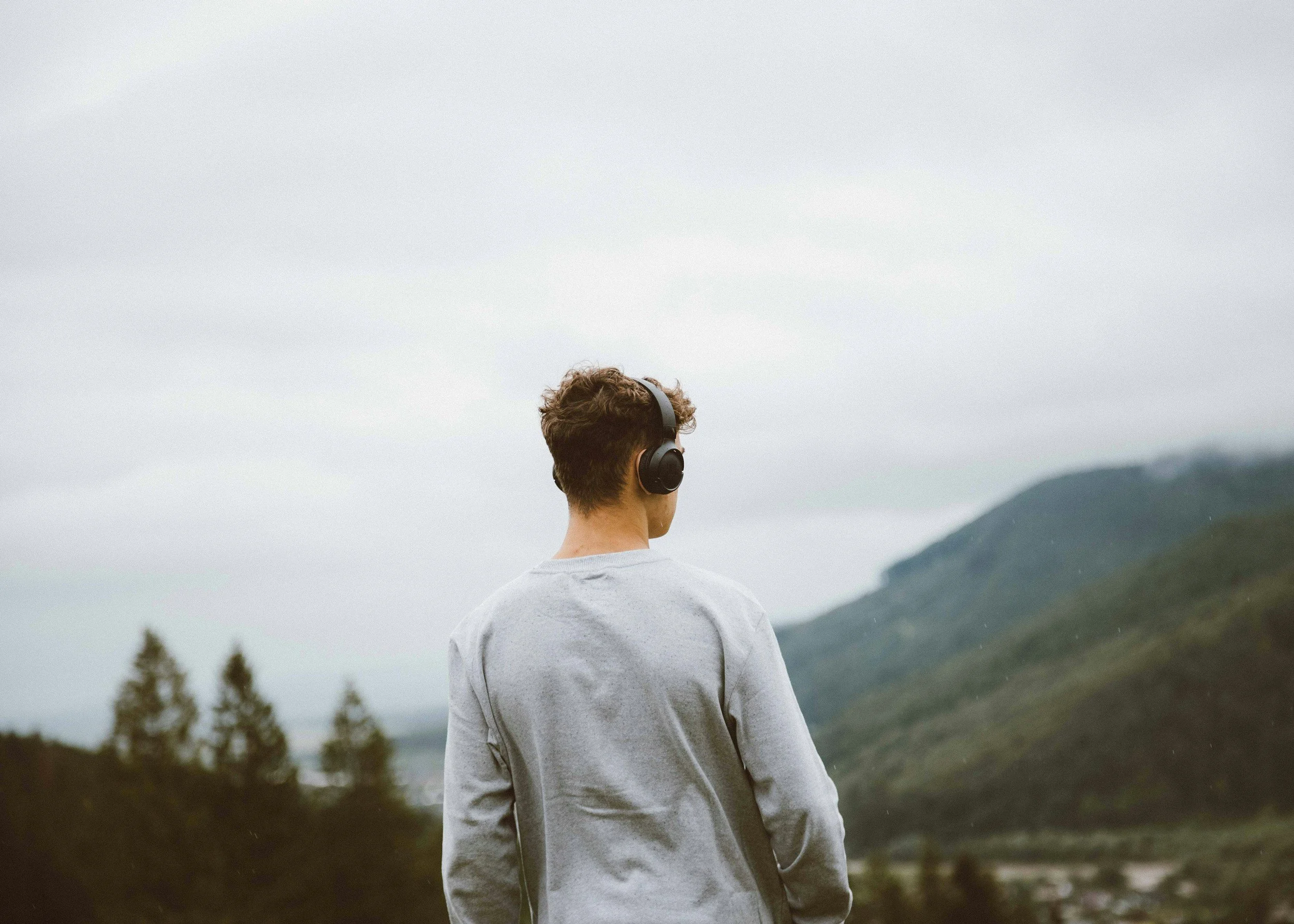 A young man with curly brown hair wearing black over-ear headphones, standing outdoors during the daytime with a cloudy sky, mountains, and trees in the background.
